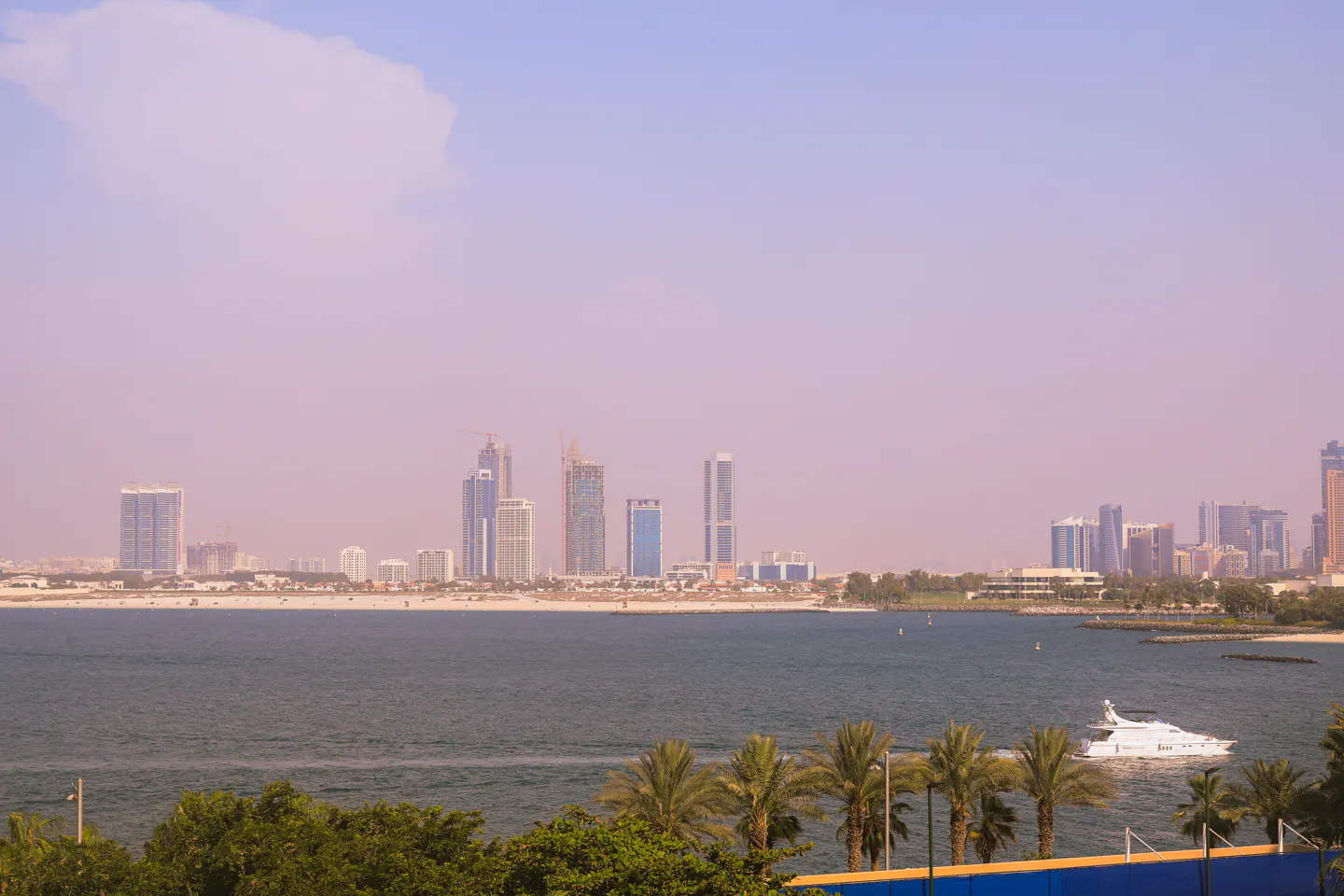 Dubai skyline view from across the water, with a white yacht, palm trees, and a blue sky.