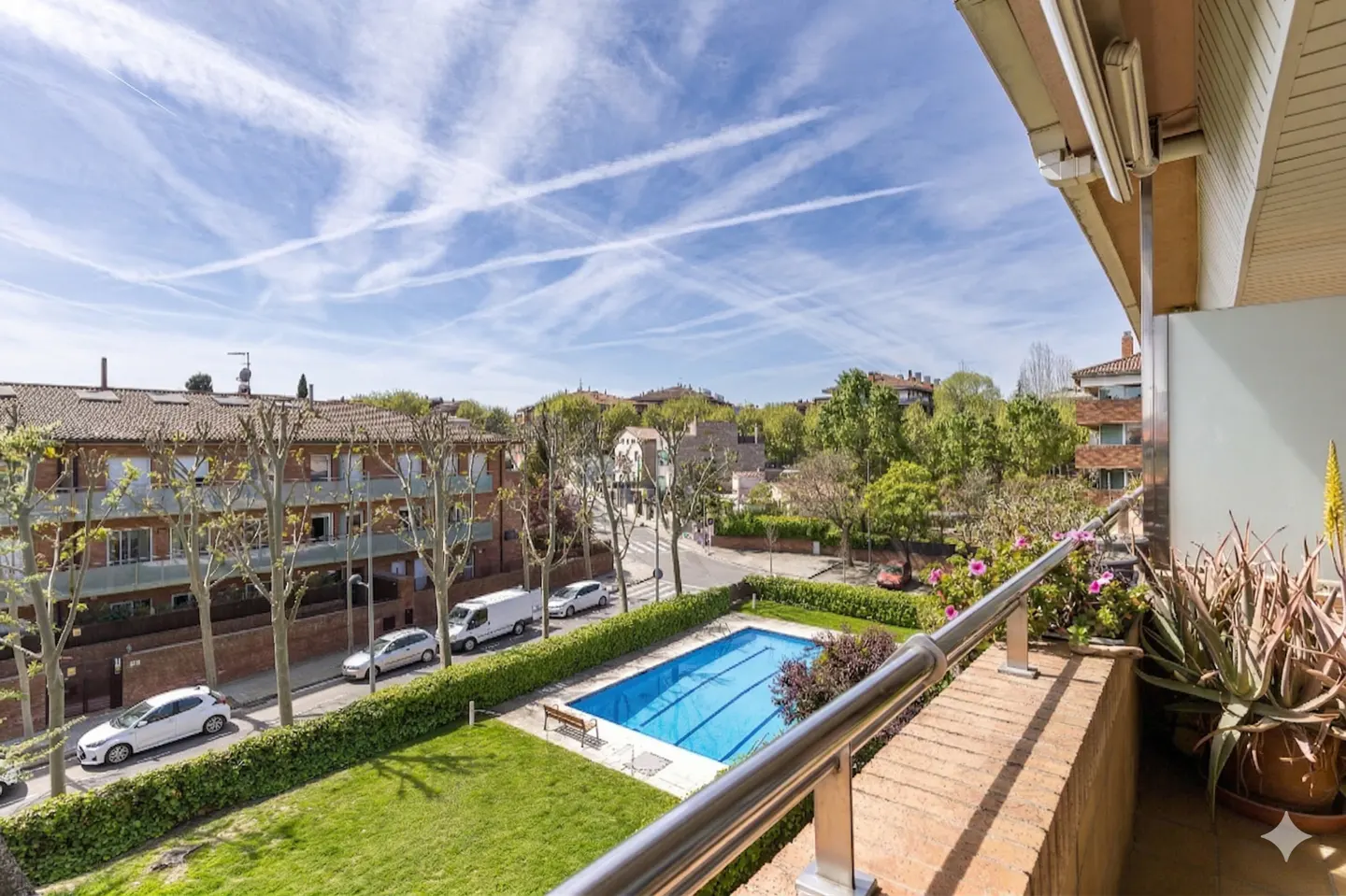 View from a balcony overlooking a blue swimming pool, green lawn, and residential street under a blue sky with contrails.
