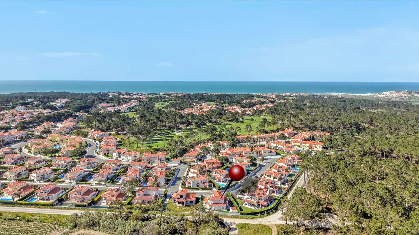 Aerial view of a coastal neighborhood with red-roofed houses, green trees, and a red pin marking a property near the ocean.
