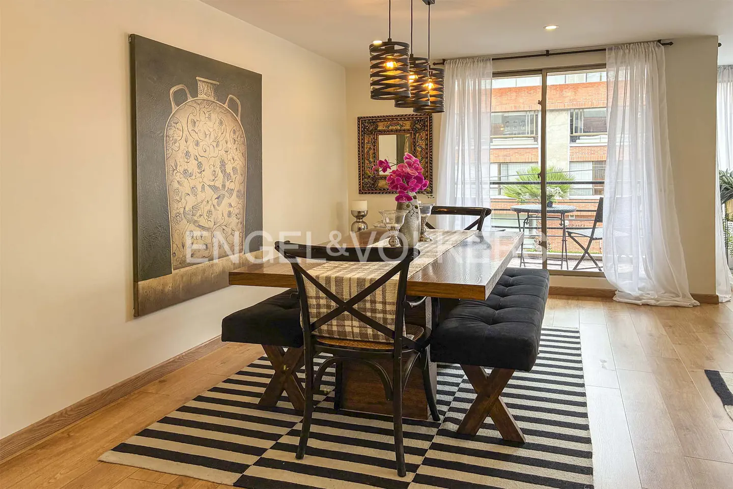 Dining room with a wooden table, black benches, and a chair on a striped rug. A painting hangs on the wall, and a balcony is visible through sheer curtains.