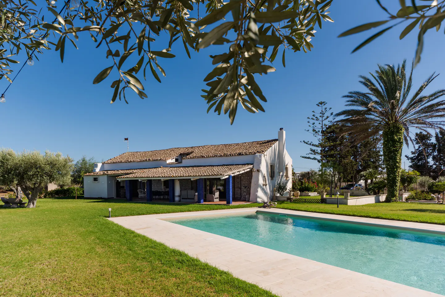 Exterior view of a white house with a brown tile roof, blue columns, a pool, and green grass.