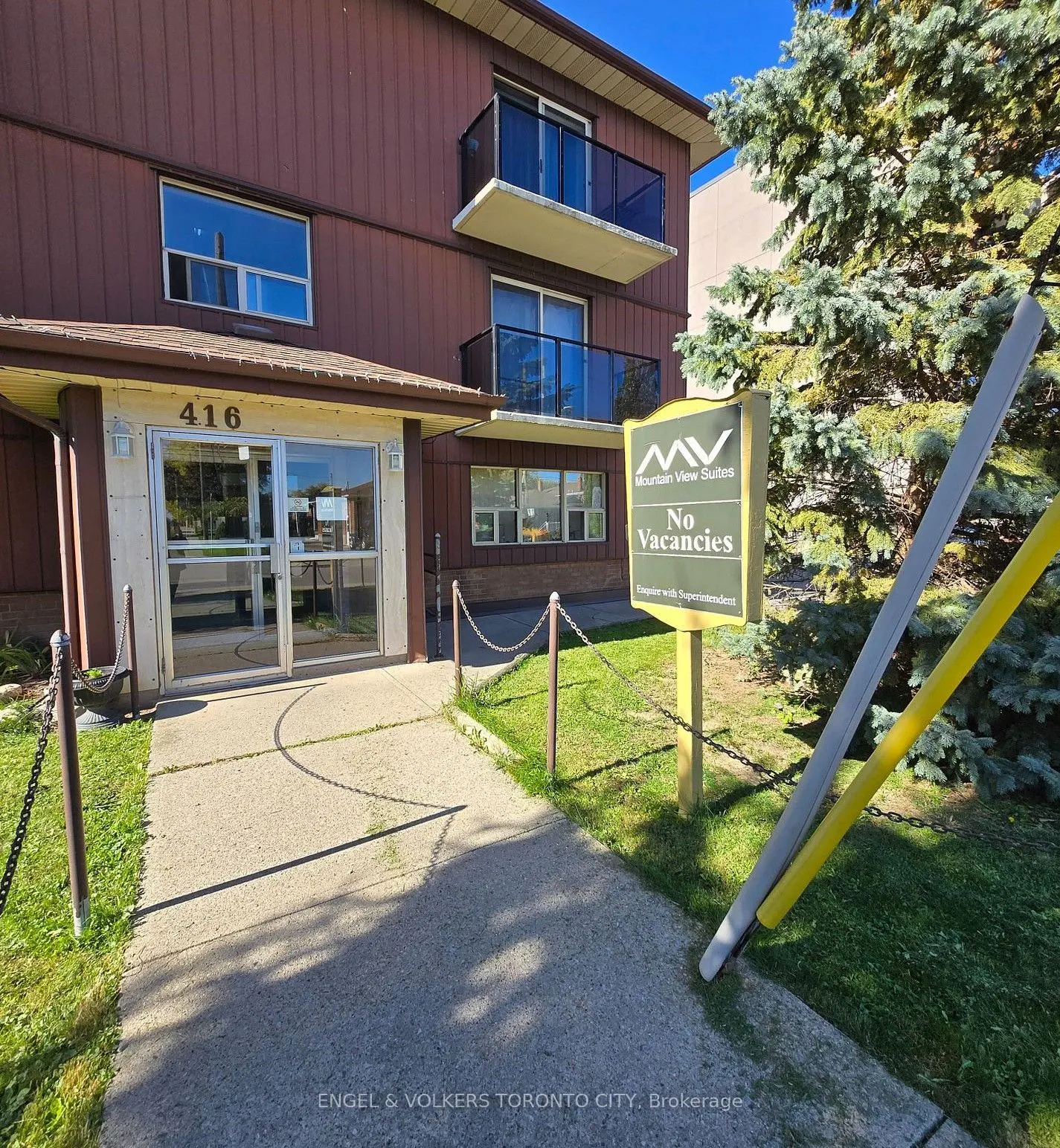 Exterior of a brown three-story apartment building with a glass entrance, balconies, and a "No Vacancies" sign on the lawn.