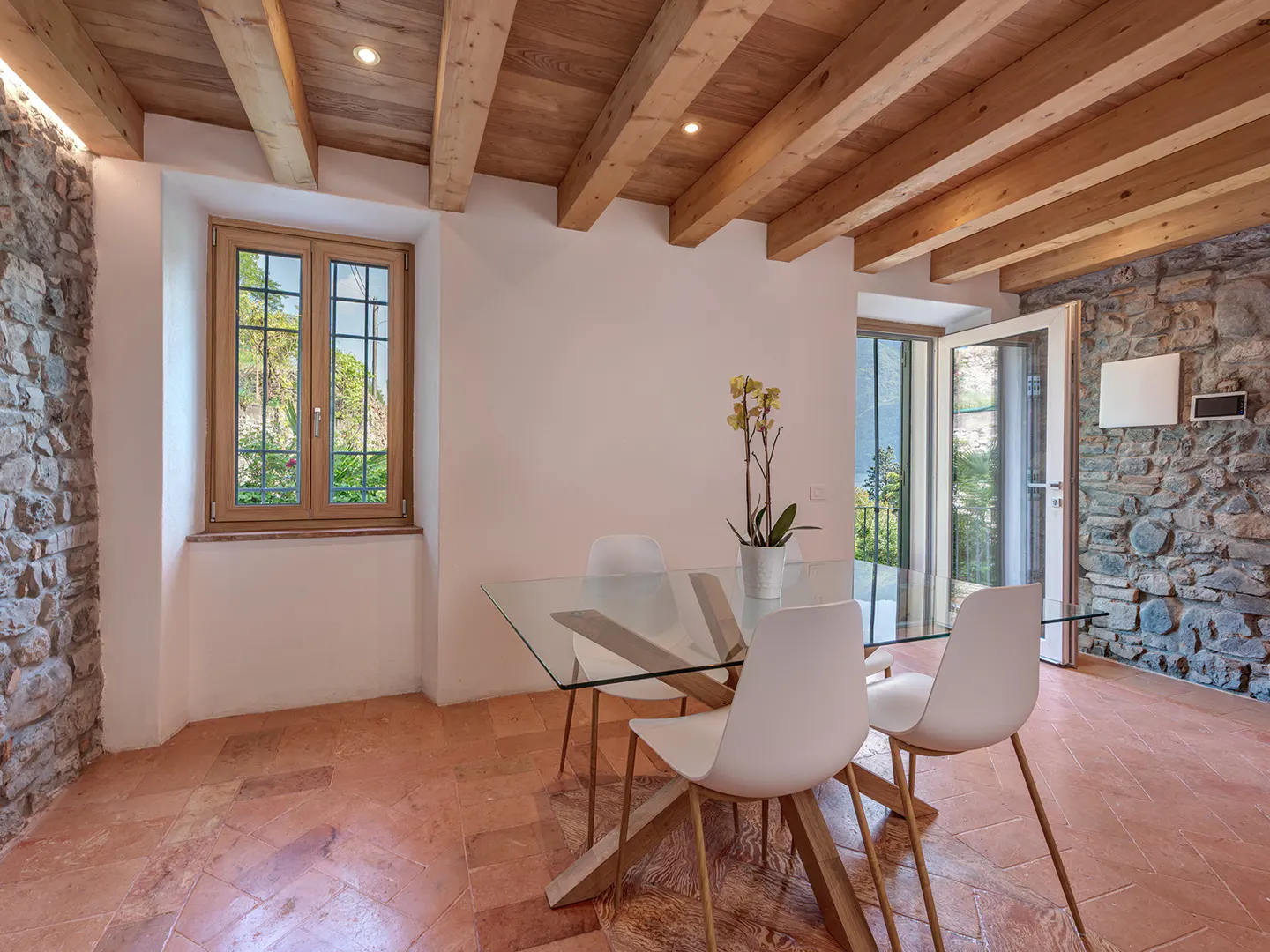 Dining room with stone walls, wood-beamed ceiling, and terracotta tile floor. A glass table with white chairs sits near a window and open doors.