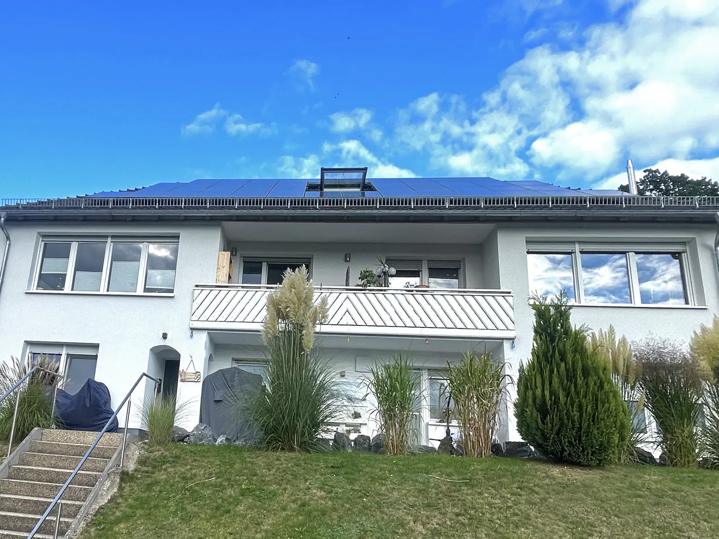 Two-story white building with solar panels on the roof, a balcony, and green lawn in front under a blue sky.