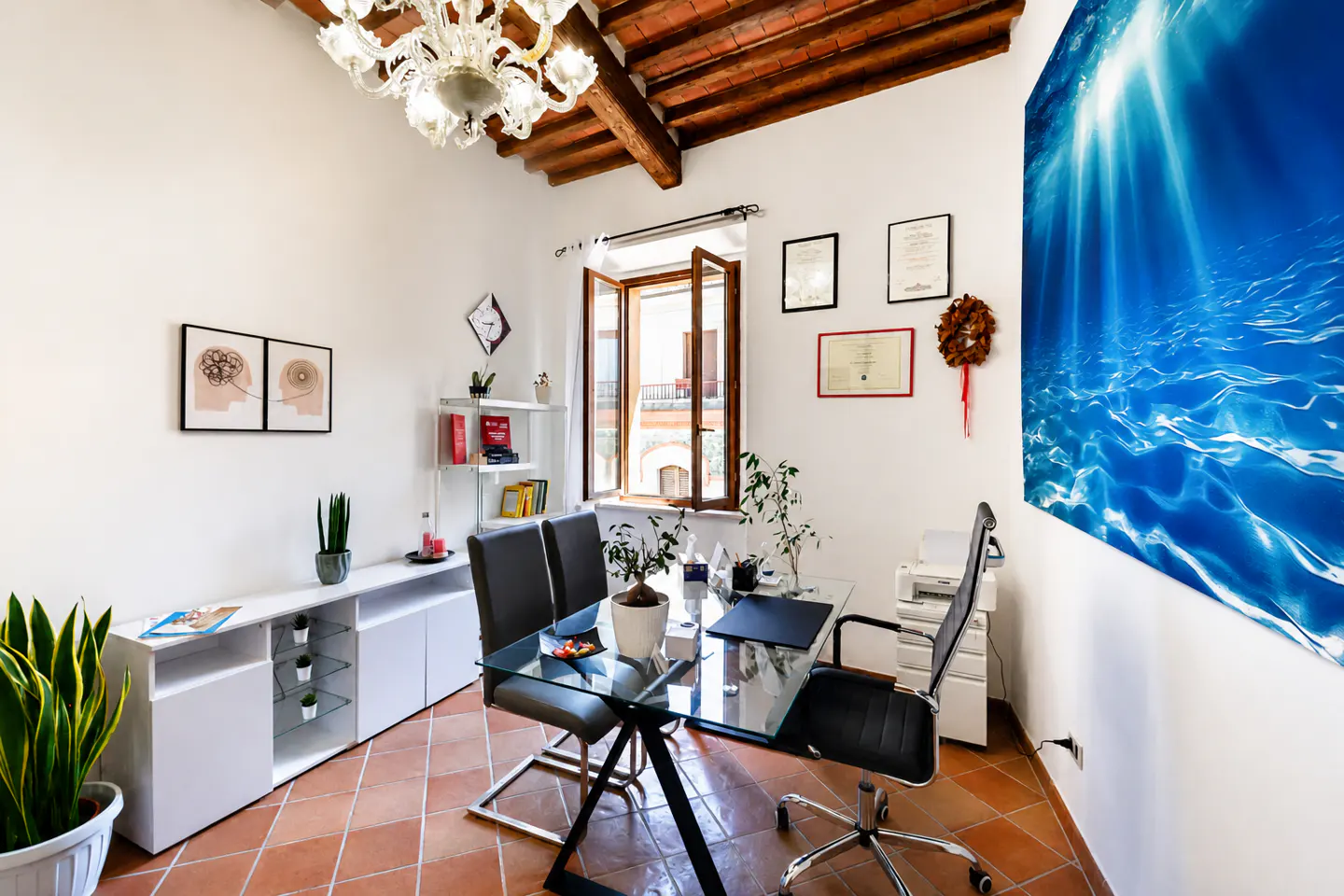 Bright office with white walls, terracotta floors, and a glass desk. A large blue ocean print hangs on the wall. A chandelier hangs from the wood-beamed ceiling.