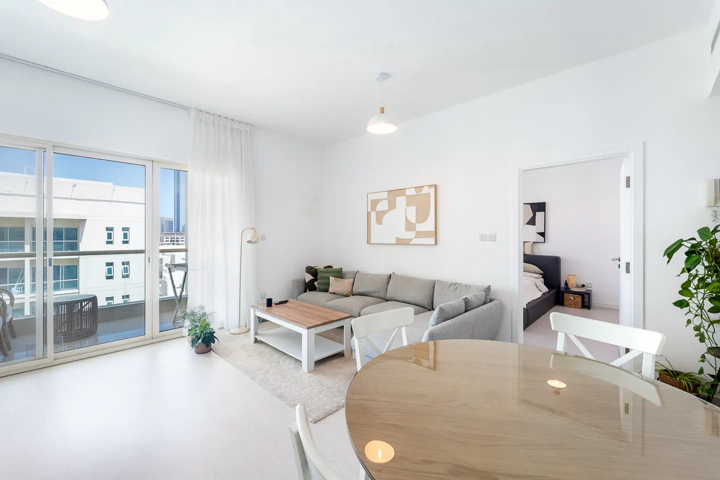 Bright, modern living room with a gray sofa, wood table, and round dining table. Balcony with city views. White walls and light wood floors.