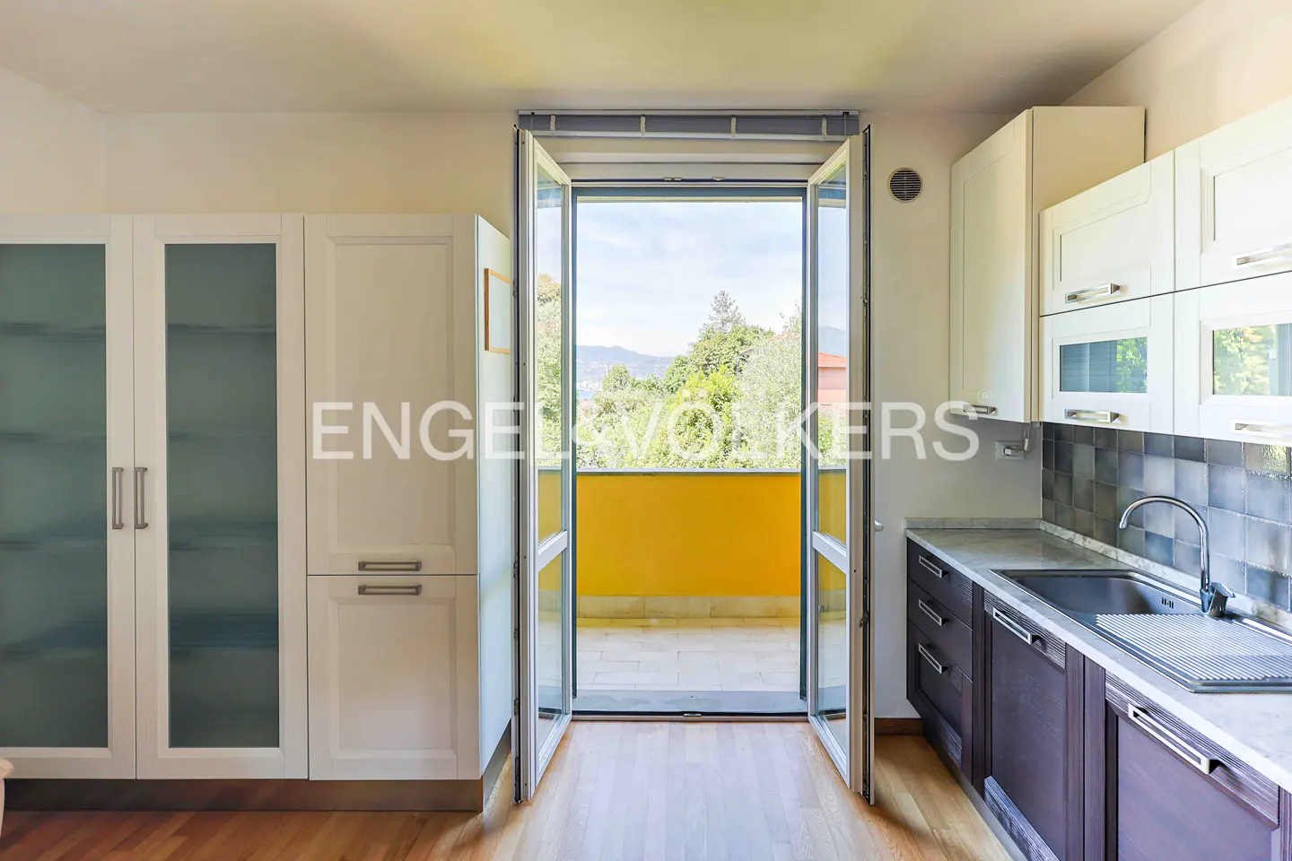 A bright kitchen with white cabinets, dark counters, and an open door to a yellow balcony with a view.
