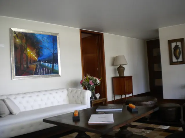 Living room with white sofa, dark wood coffee table, and painting of a river at night. A lamp and flowers sit on a side table.