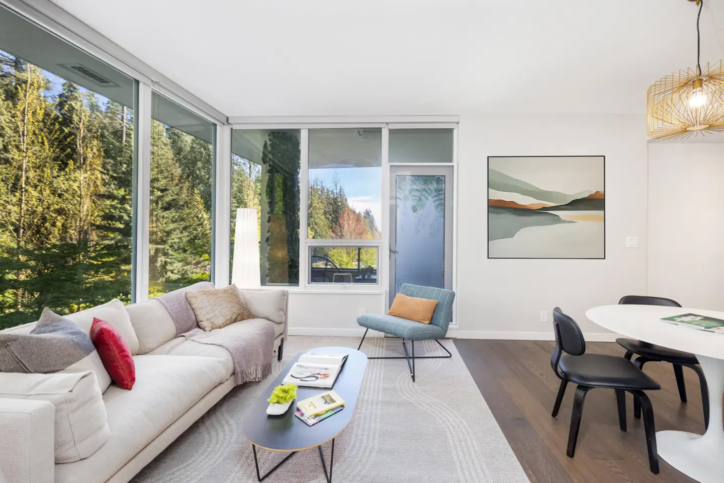 Bright living room with a white sofa, blue chair, and black coffee table on a patterned rug. Large windows overlook a forest. A white table and black chairs are in the dining area.