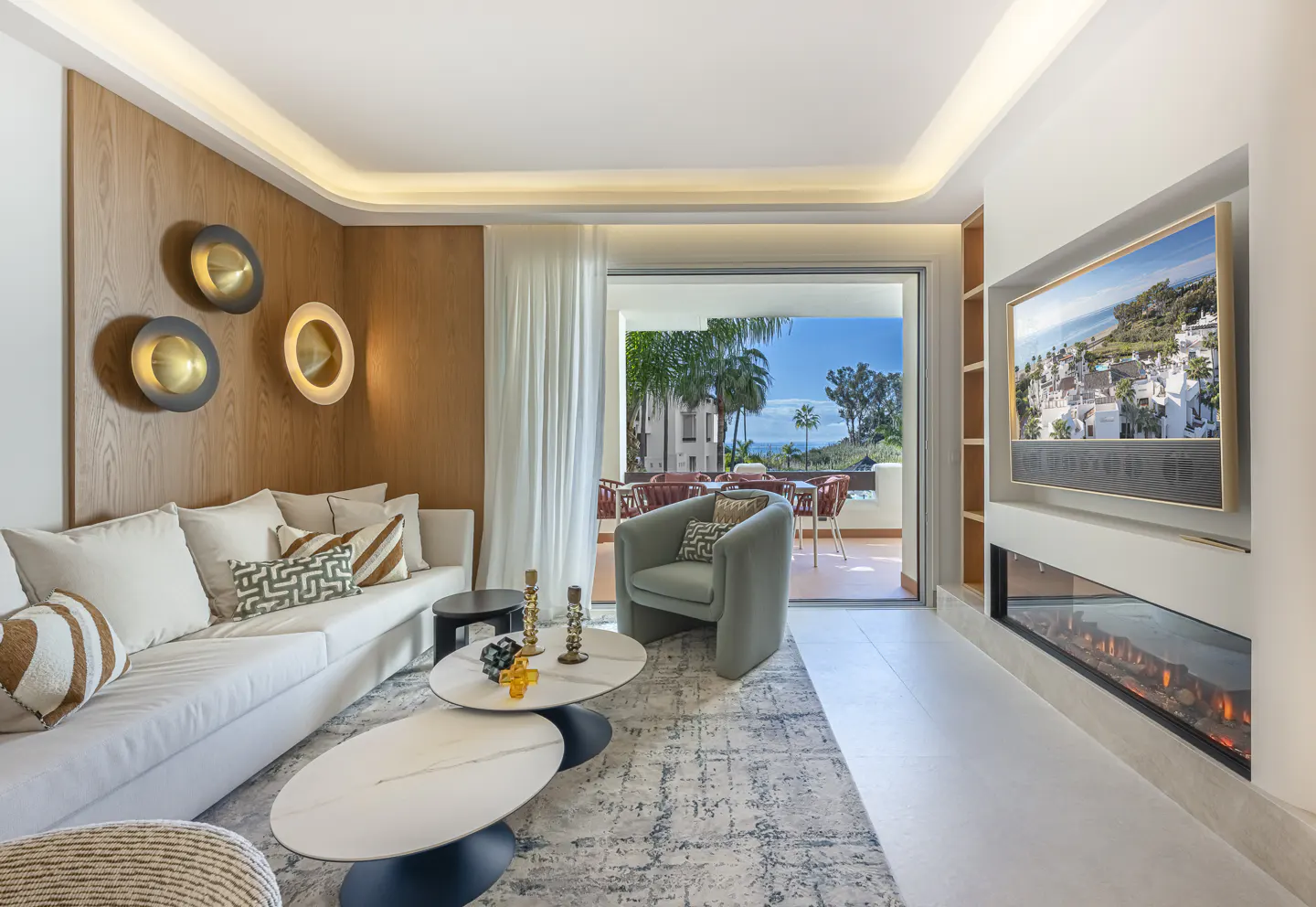 Bright living room with a white sofa, blue chair, and a view of a balcony with palm trees and the ocean. A modern fireplace is visible.