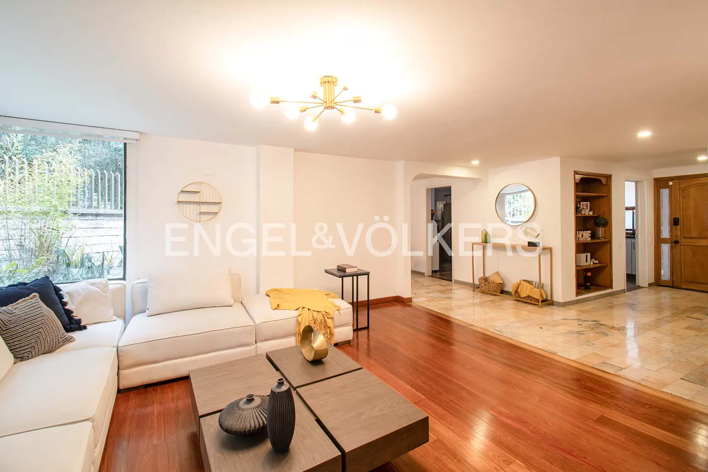 Bright living room with a white sectional sofa, dark wood coffee table, and hardwood floors. Engel & Völkers logo on the wall.