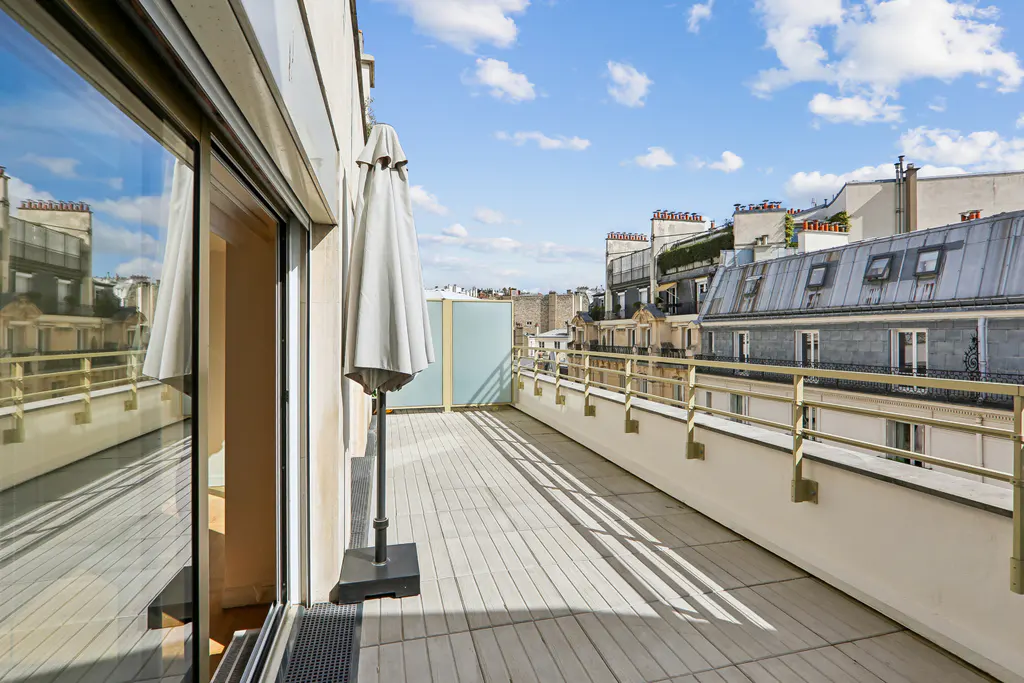 Wide balcony with gray tiles, white railings, and a closed umbrella. City buildings are visible in the background under a blue sky with clouds.