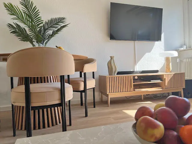 Living room with a TV, wooden cabinet, and round table with two beige chairs. A bowl of fruit sits on a table in the foreground.