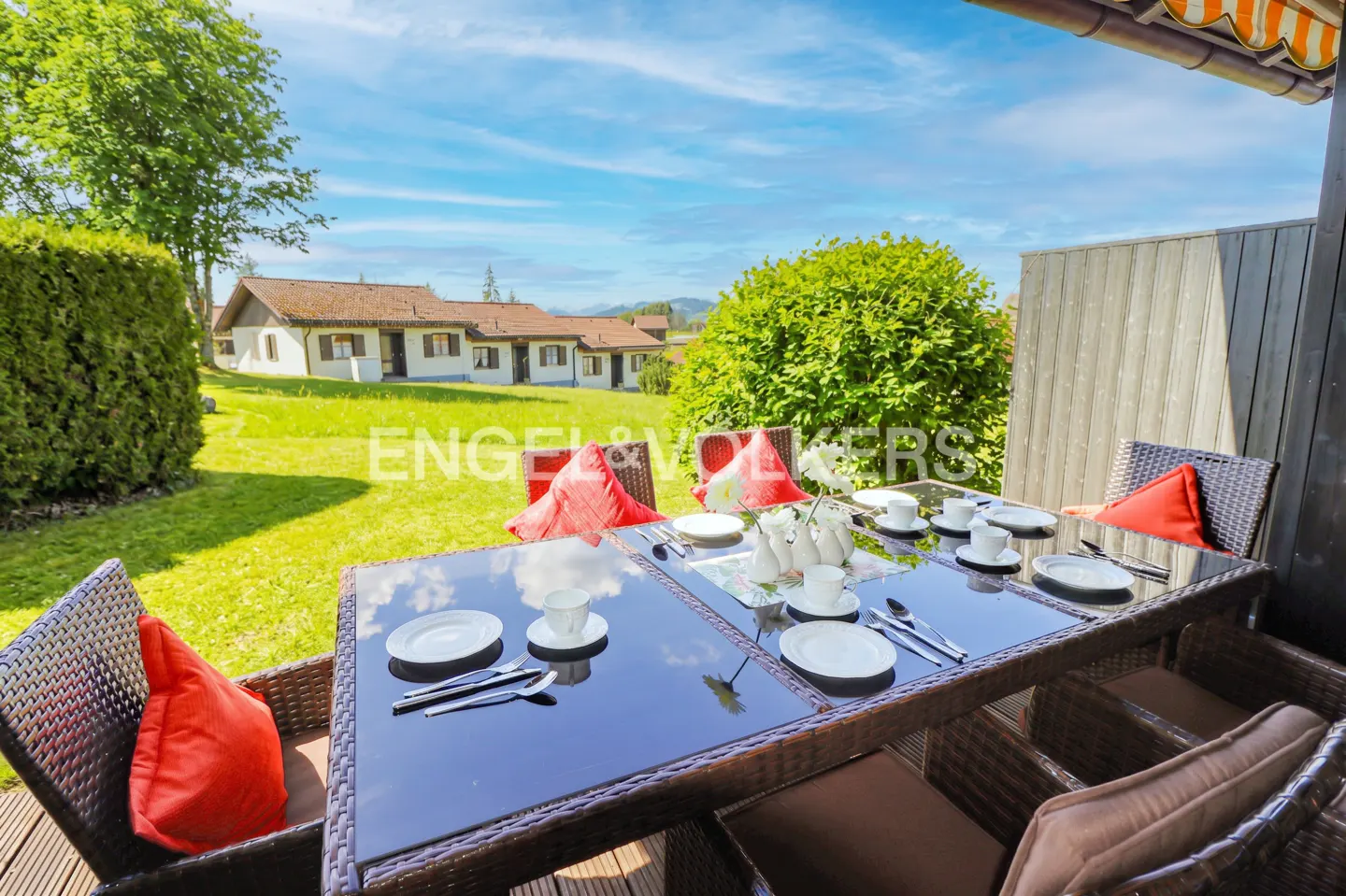 Outdoor patio with a set table, wicker chairs, and red pillows. A green lawn and houses are in the background.