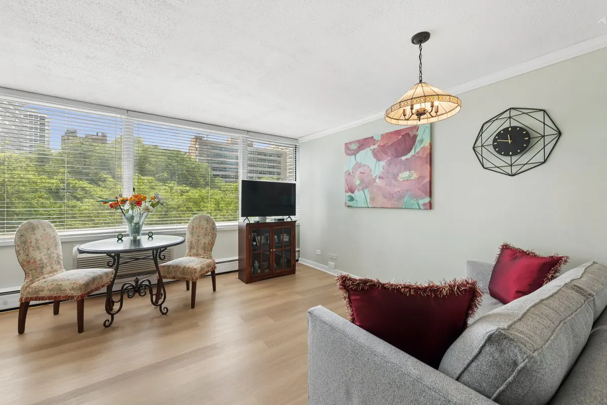 Living room with wood floors, a gray sofa with red pillows, and a table with two chairs by a window overlooking trees.