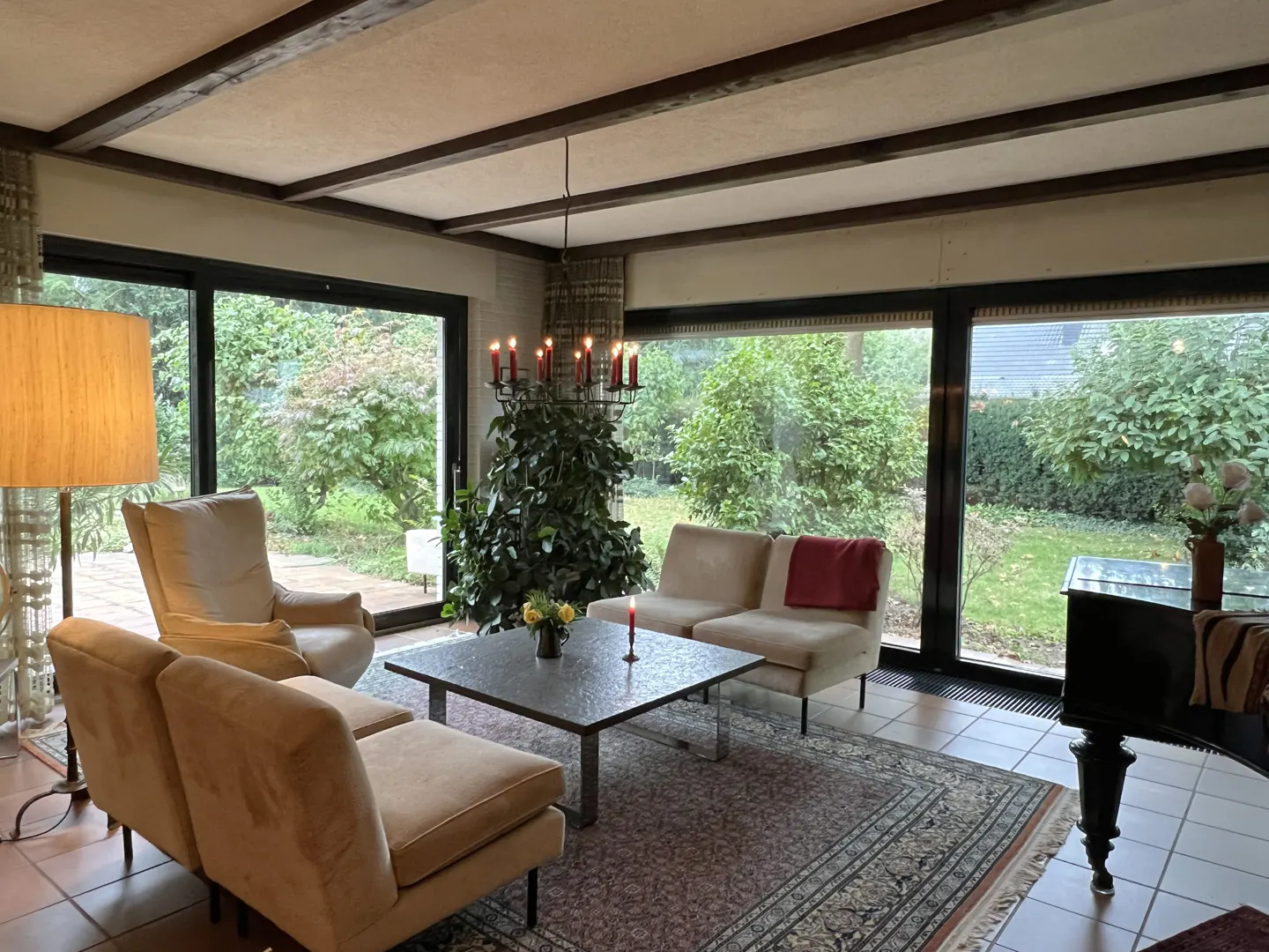 Living room with beige furniture, a black piano, and large windows overlooking a green garden.