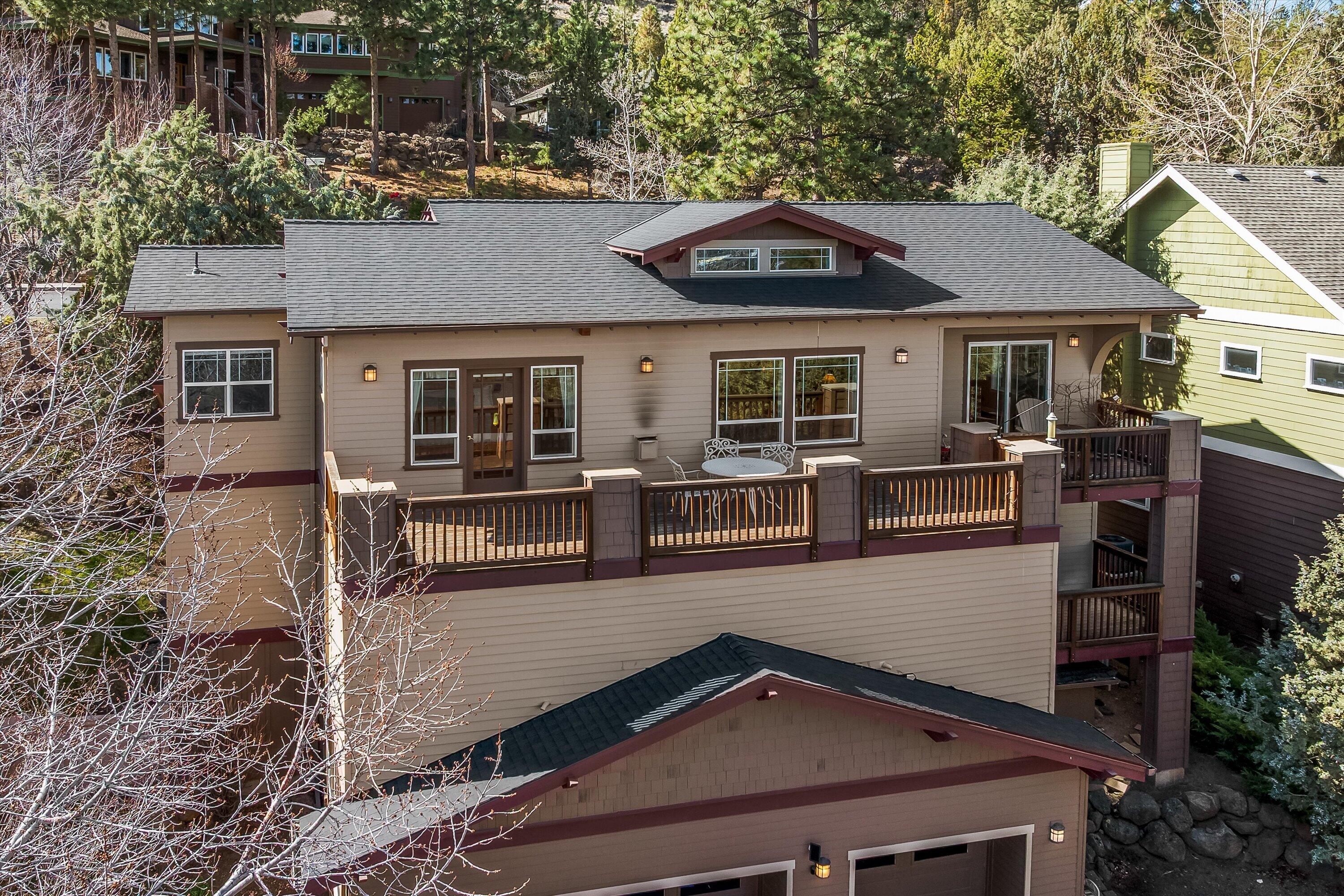 Tan two-story house with a dark roof, brown trim, and a wooden deck with a white table and chairs. The house is surrounded by trees.