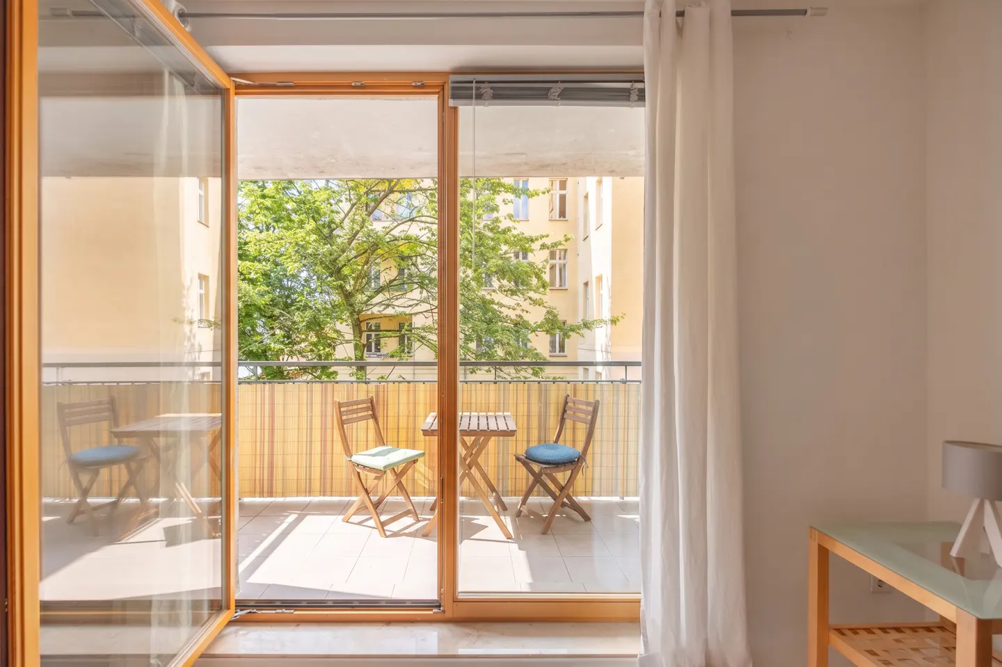 Open glass doors lead to a balcony with a table and two chairs. Trees and a building are visible in the background.