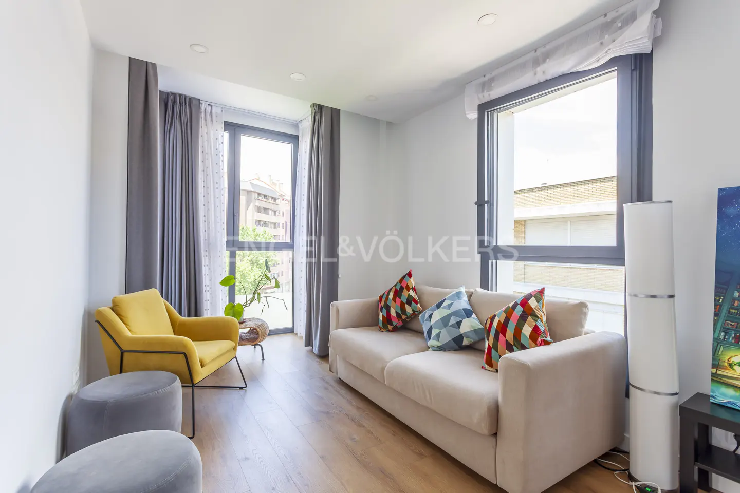Bright living room with a beige sofa, yellow armchair, two grey ottomans, and large windows with grey curtains.