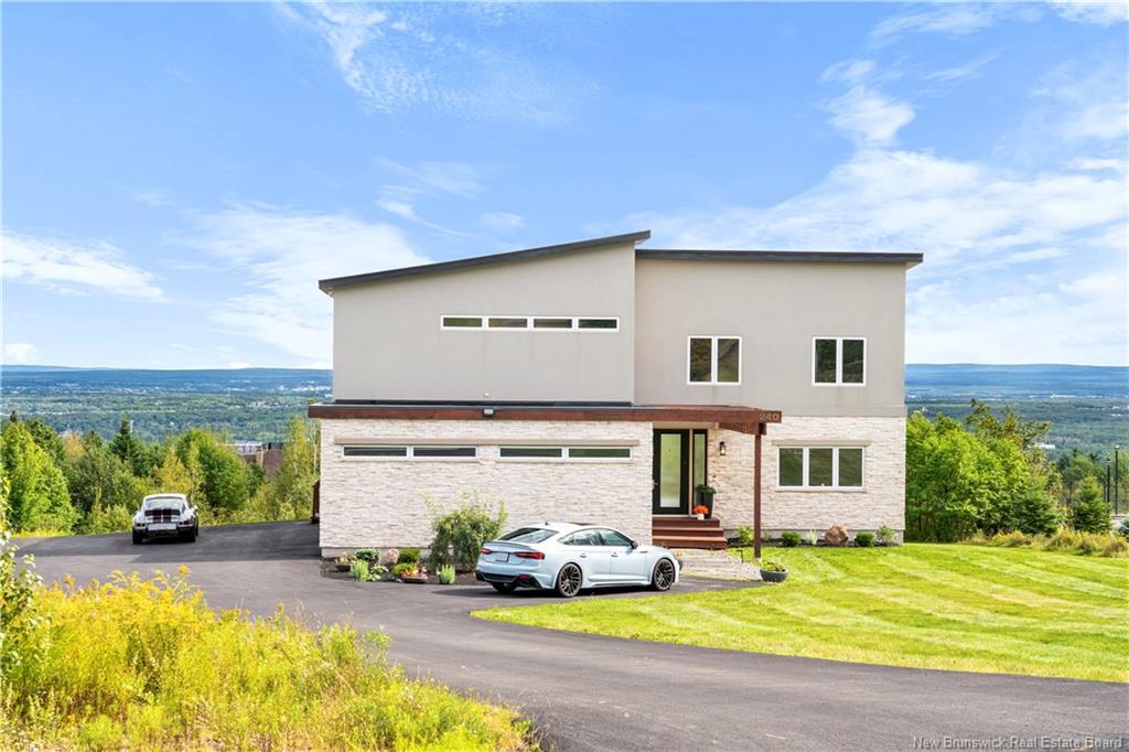 Modern two-story home with stone and grey siding, a paved driveway, two cars, and a scenic valley view under a blue sky. [AI-generated text]