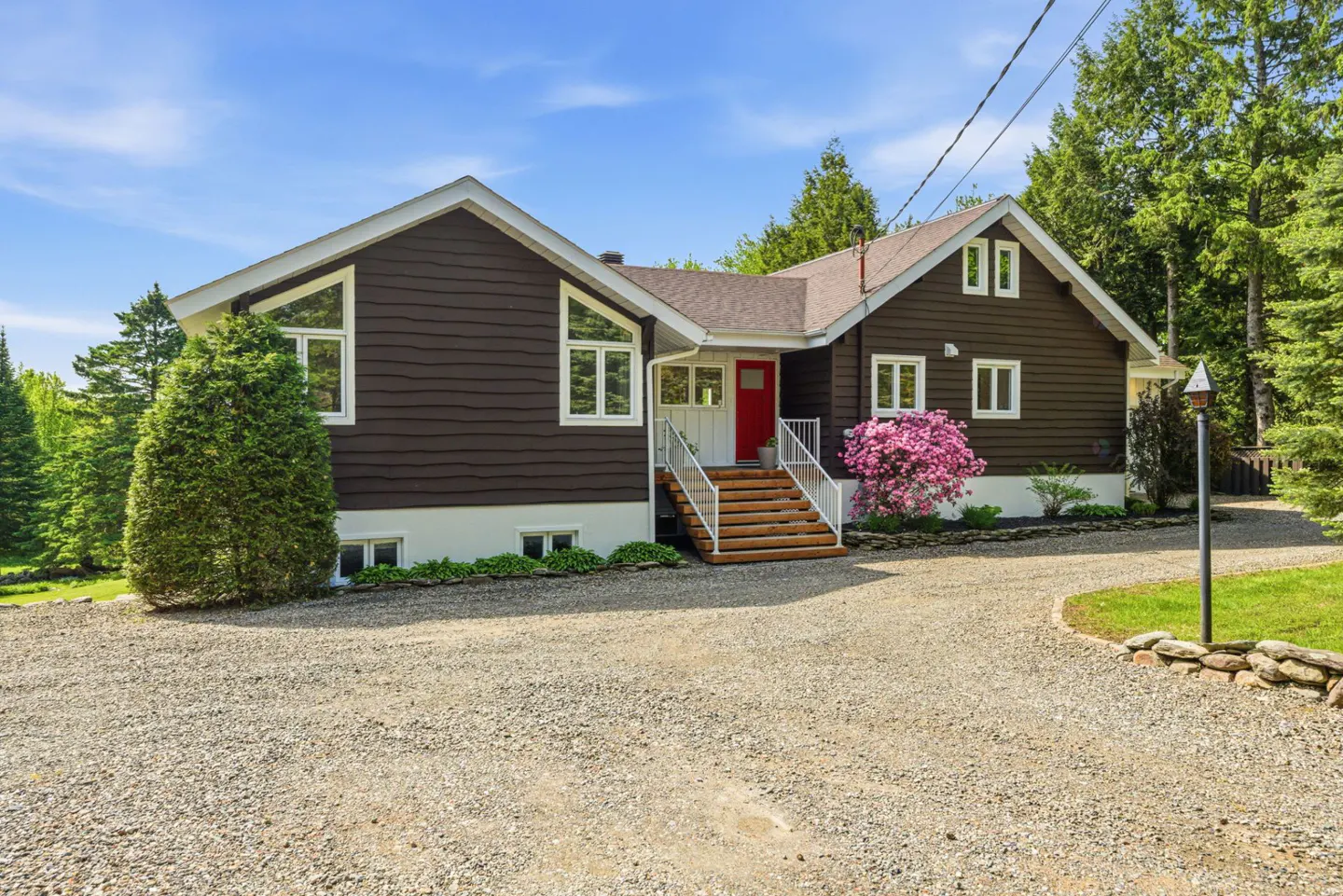 A brown house with white trim and a red door sits on a gravel driveway with green trees in the background.