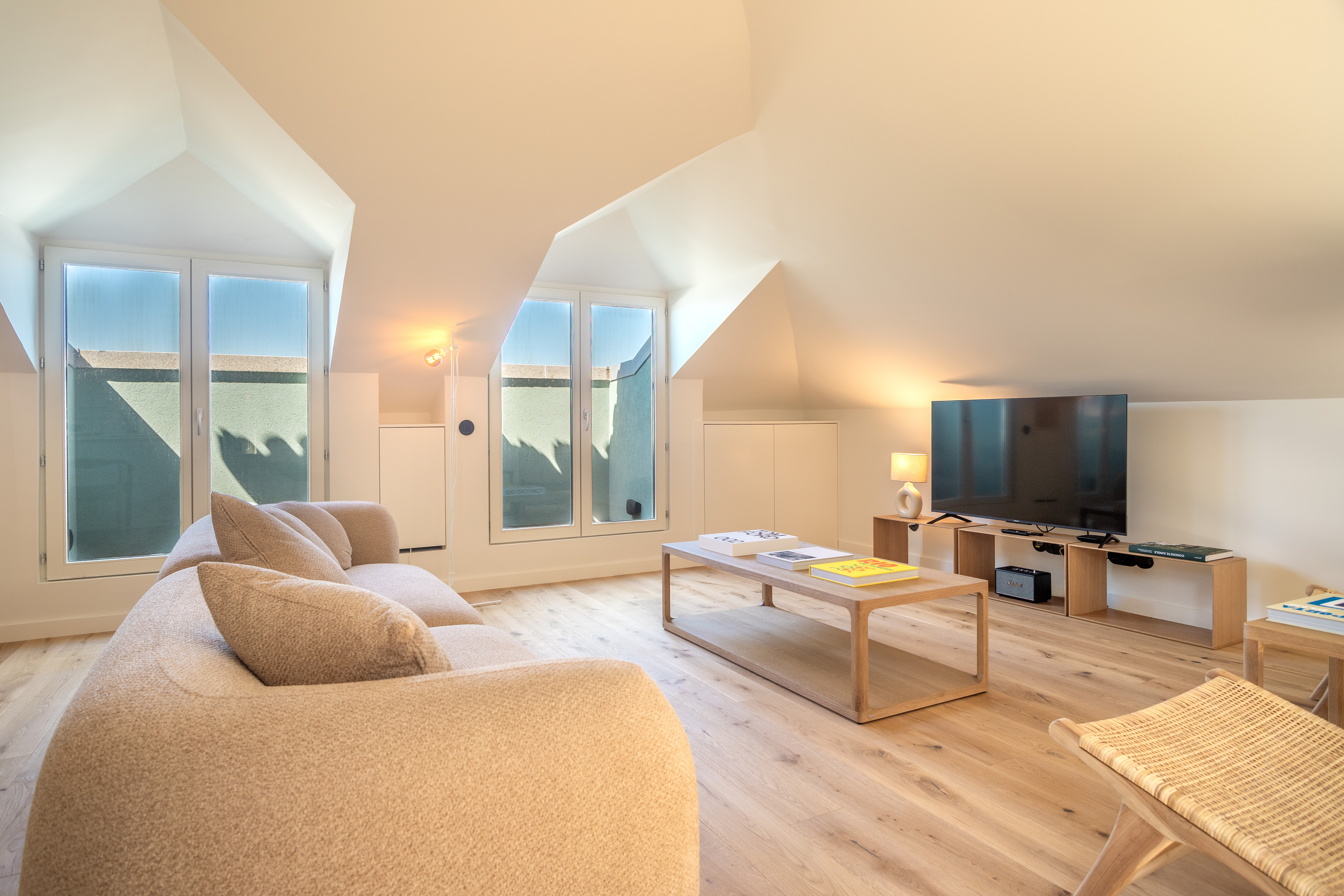 Bright attic living room with a beige sofa, wooden coffee table, TV on a low stand, and large windows with natural light. [AI-generated text]