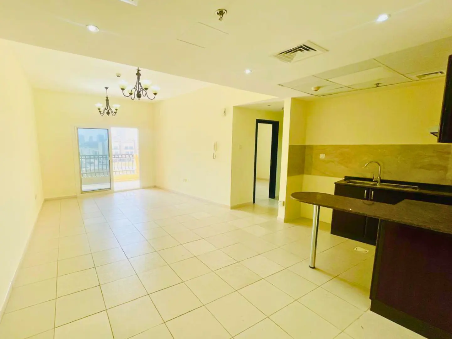 Bright, empty apartment with tile floors, chandeliers, and a balcony. A kitchen area with dark cabinets is visible.
