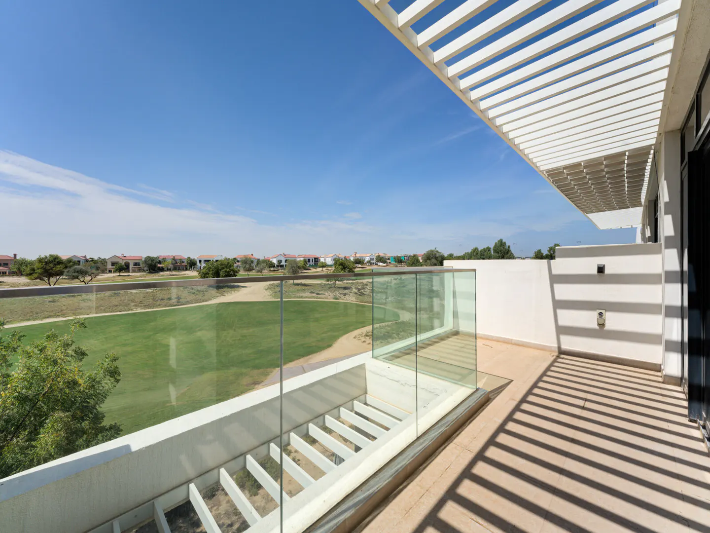 Balcony view of a green golf course under a blue sky. White pergola casts shadows on the beige tile floor. Glass railing.