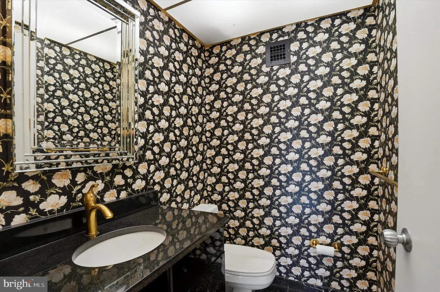 A bathroom with black floral wallpaper, a black granite countertop, and a gold faucet. A toilet and a mirror are also visible.