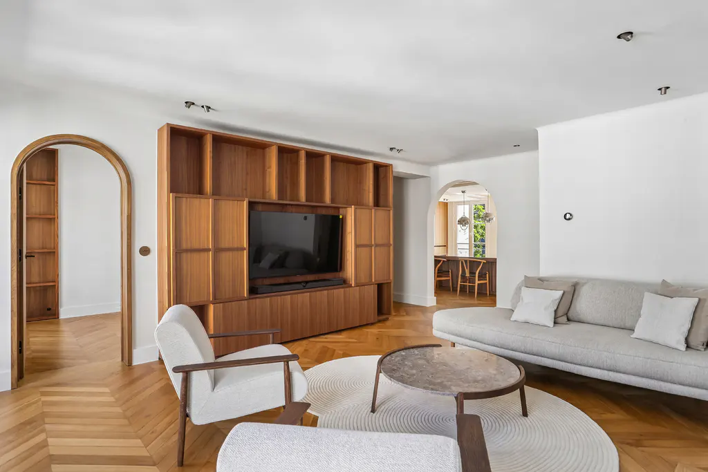 Modern living room with herringbone wood floors, a large wooden media unit, a grey sofa, and two white armchairs.