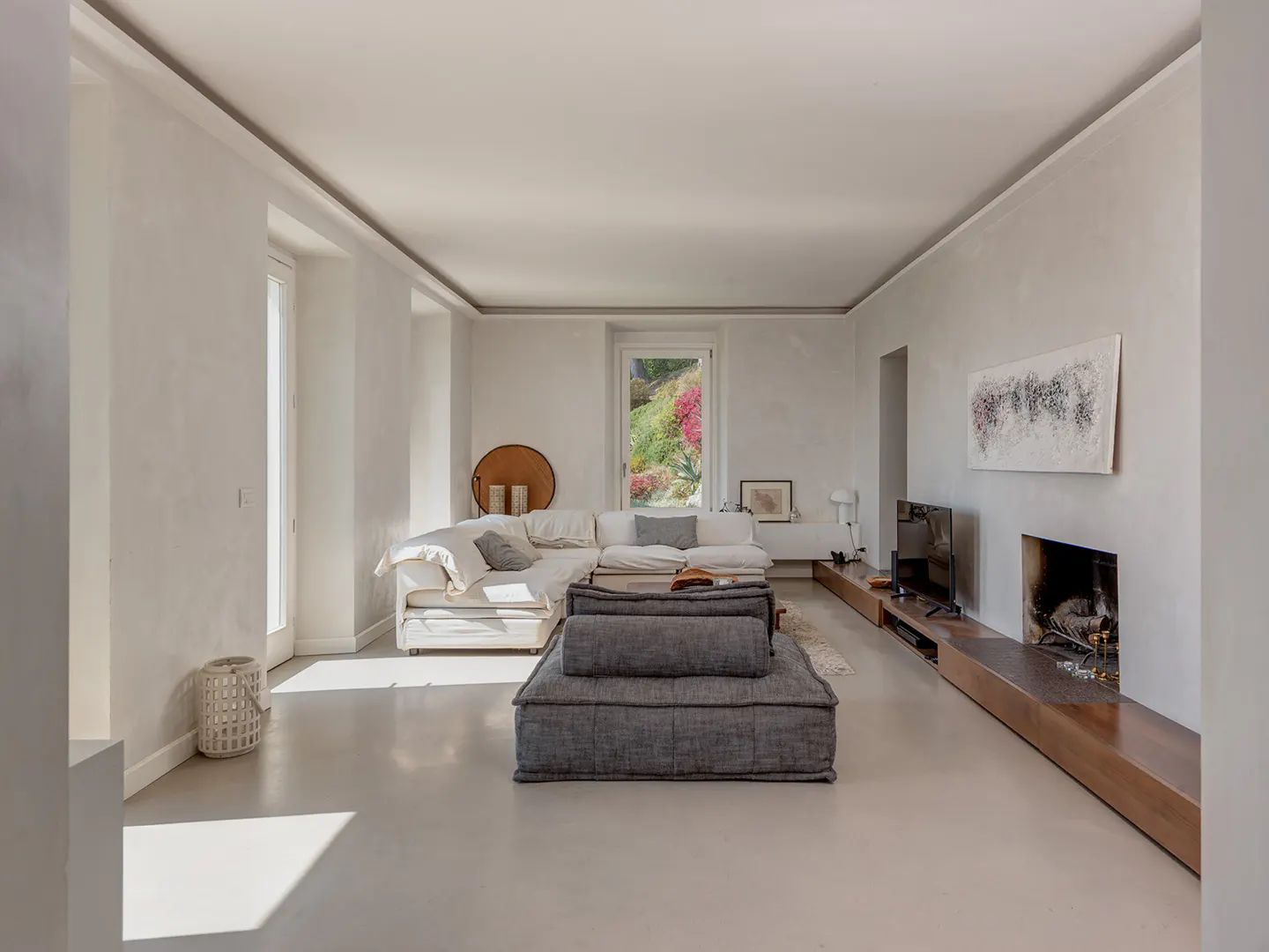 Bright, modern living room with white walls, gray sofa, and a fireplace. A window shows a lush green landscape.