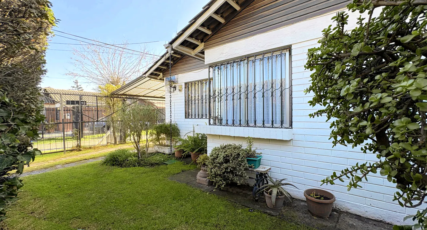 Exterior view of a white brick house with barred windows, green lawn, and various potted plants.