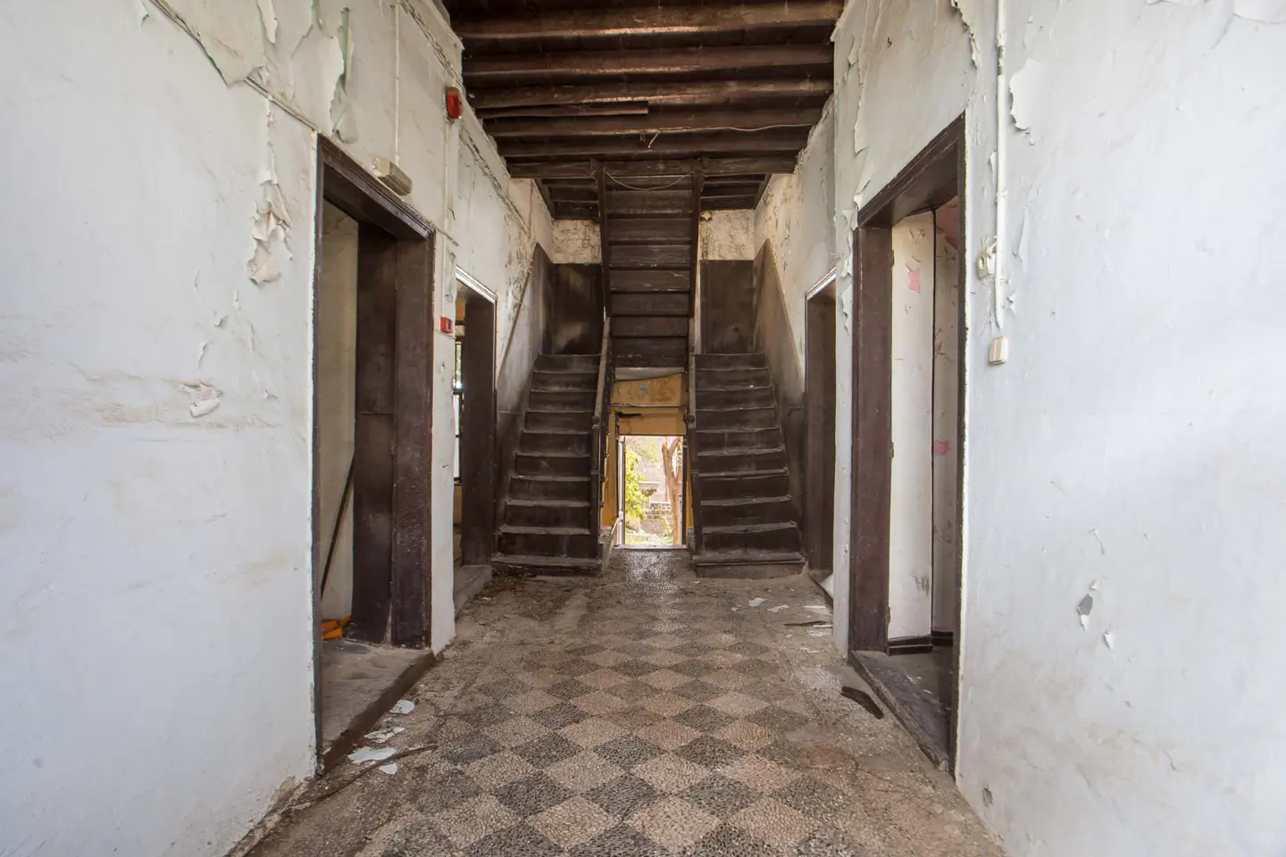 Hallway with peeling white walls, brown doors, and checkered tile floor. Two staircases lead up to a dark wooden ceiling.