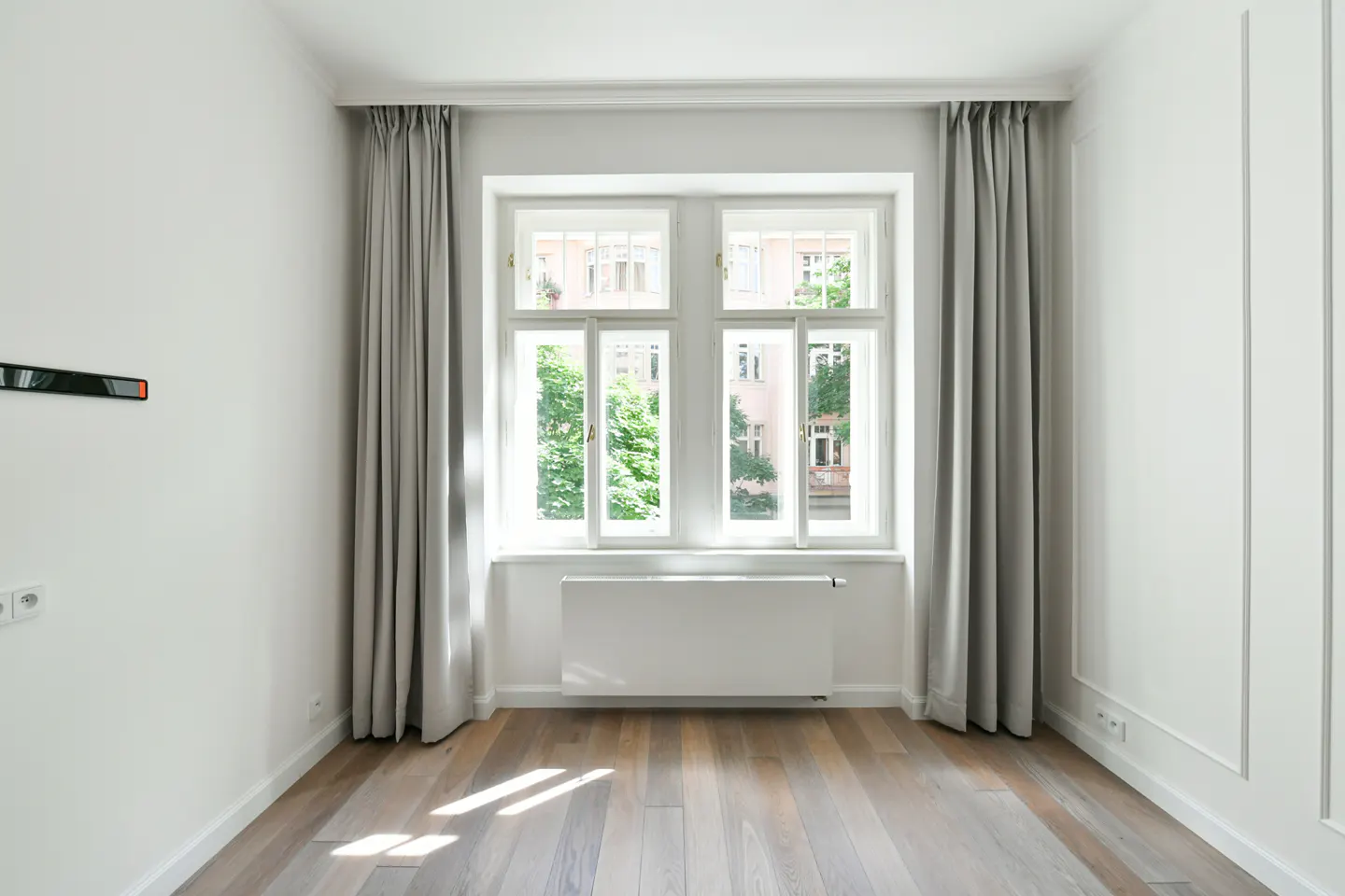 Bright, empty room with hardwood floors, white walls, and a large window with gray curtains. Sunlight streams in, illuminating the space.