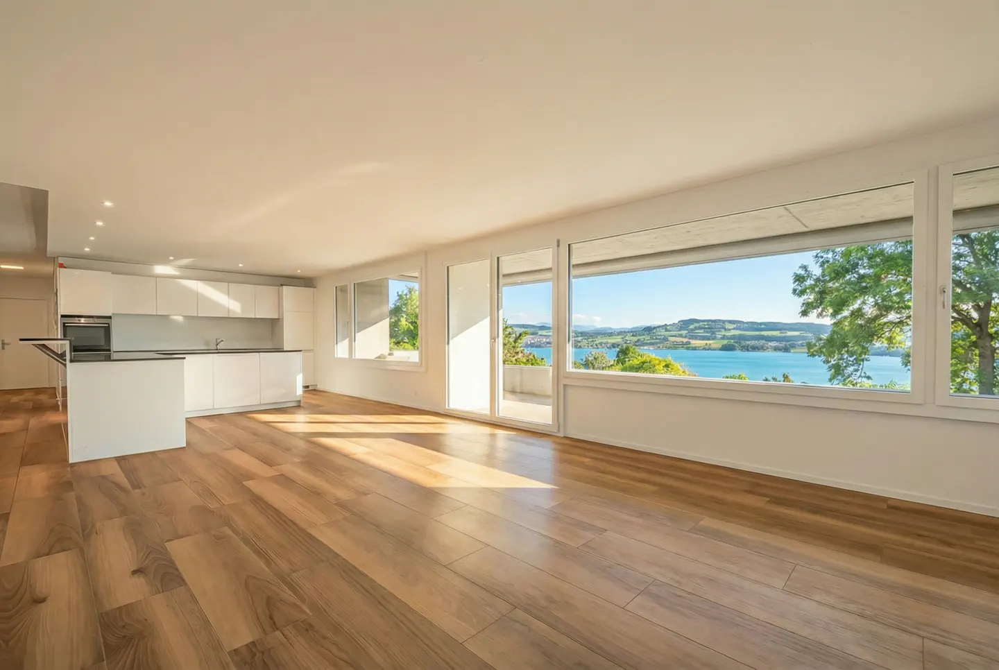 Bright, modern apartment interior with wood floors, white kitchen, and large windows overlooking a lake.