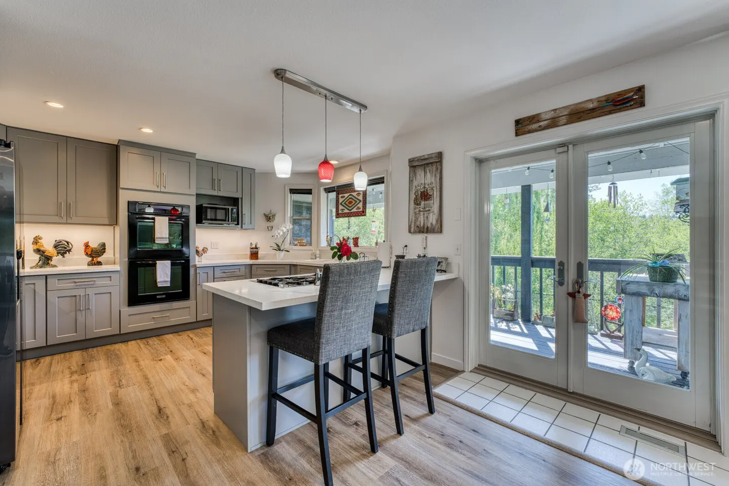Bright kitchen with gray cabinets, wood floors, and a white island with two gray chairs. French doors lead to a deck with green trees in the background.
