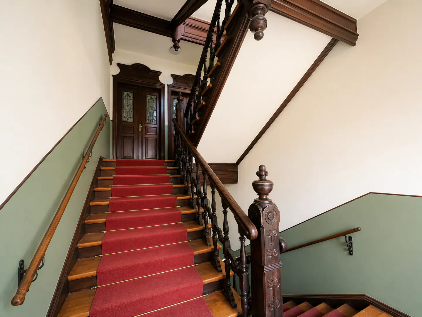 Grand staircase with a red carpet runner, dark wood banisters, and sage green walls leading to ornate wooden doors.