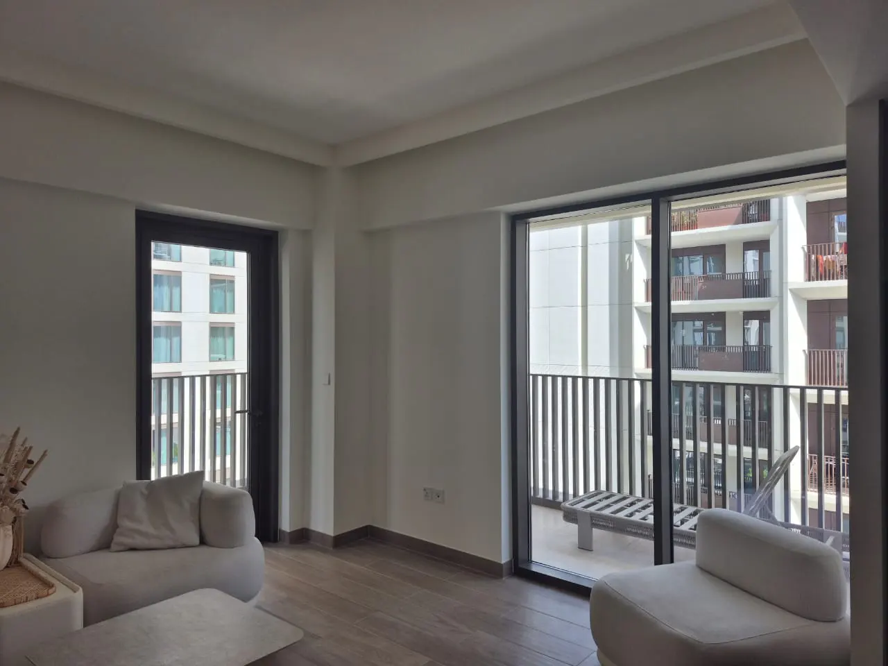 Bright living room with white walls, wood floors, and modern furniture. Balcony with black railings overlooks a building. Door to another balcony on the left.
