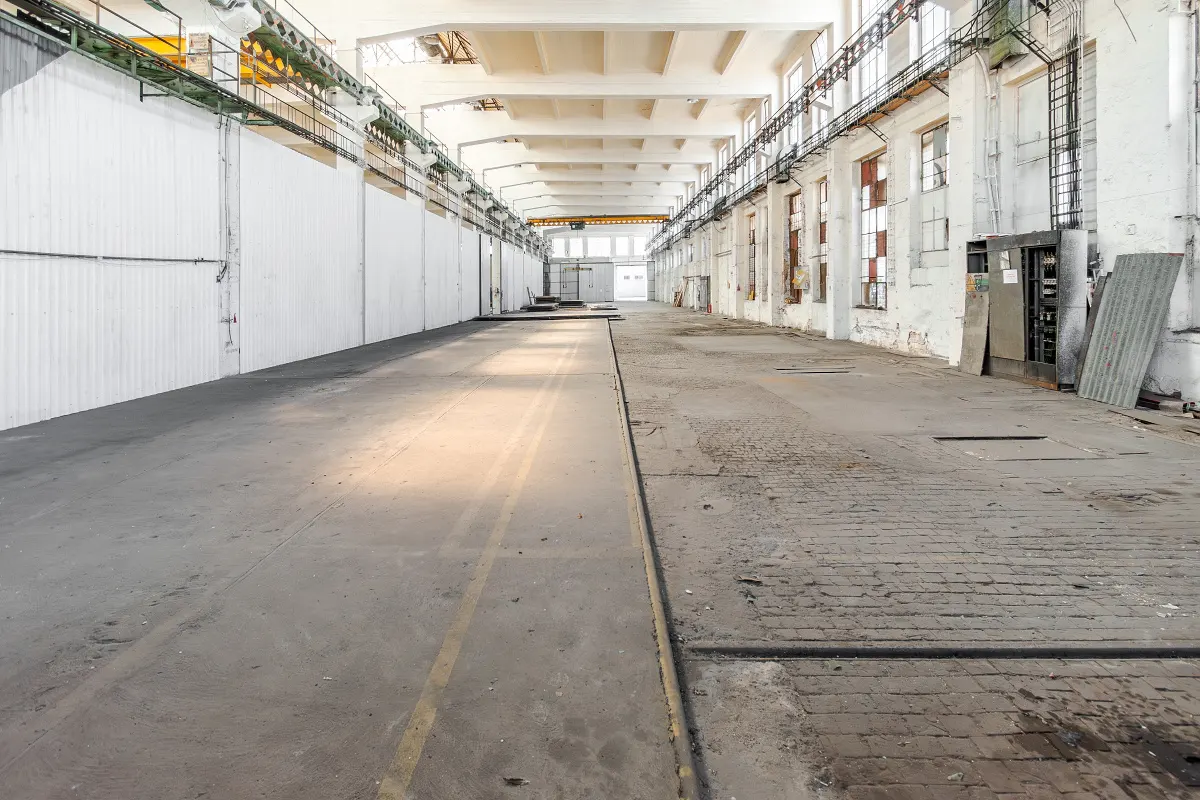 A long, empty warehouse with white walls and a concrete floor. Windows line one side, and a crane hangs from the ceiling.