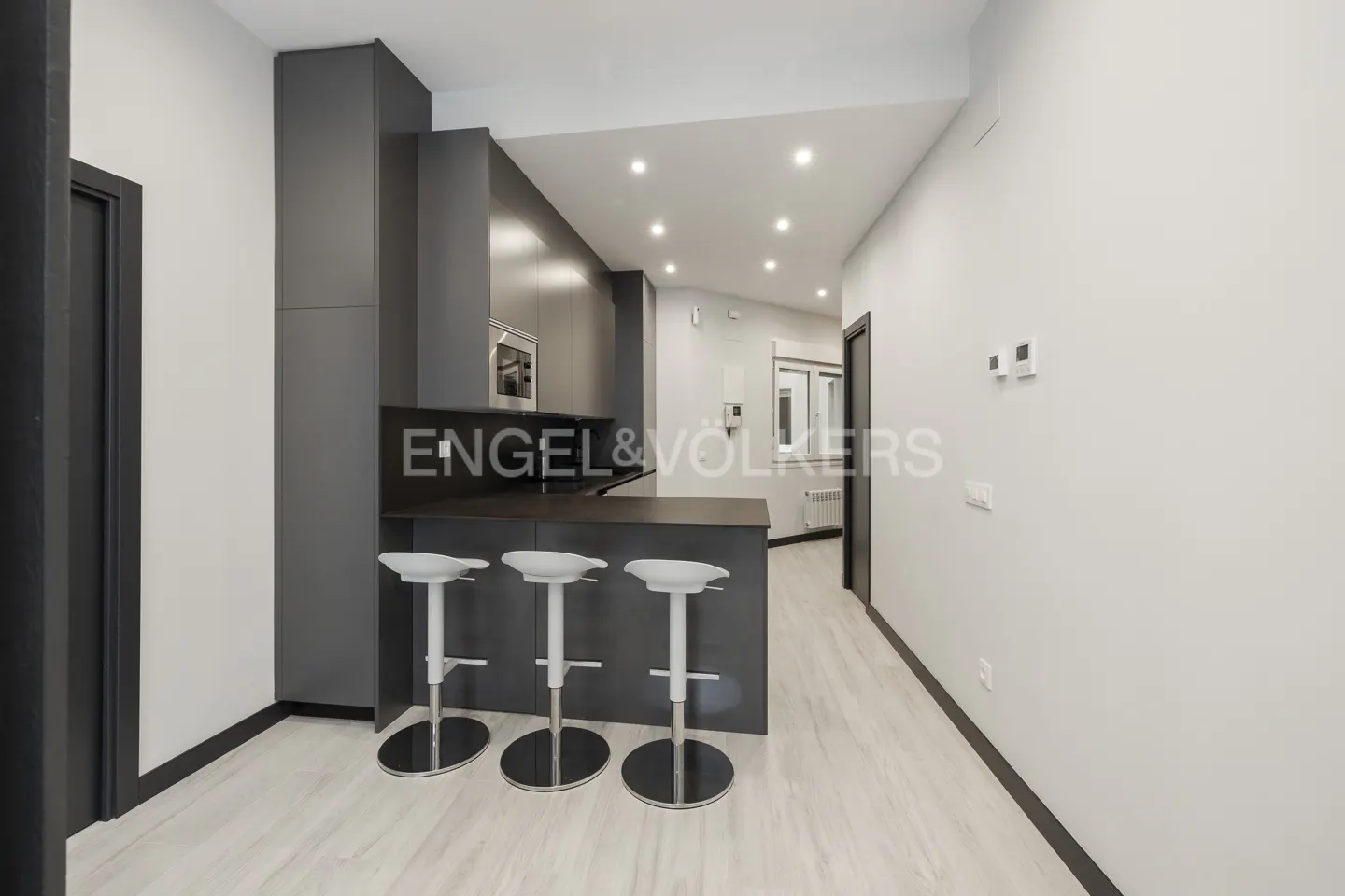 A modern kitchen with gray cabinets, a breakfast bar with three white stools, and light wood floors. The Engel & Volkers logo is visible.