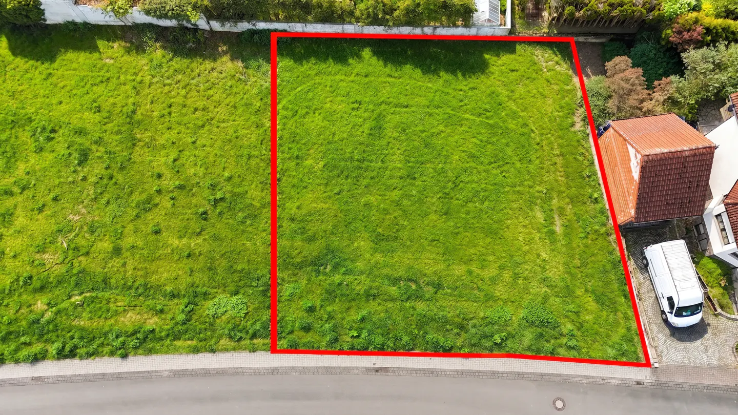 Aerial view of a vacant lot with green grass, outlined in red, next to a house and a white van.
