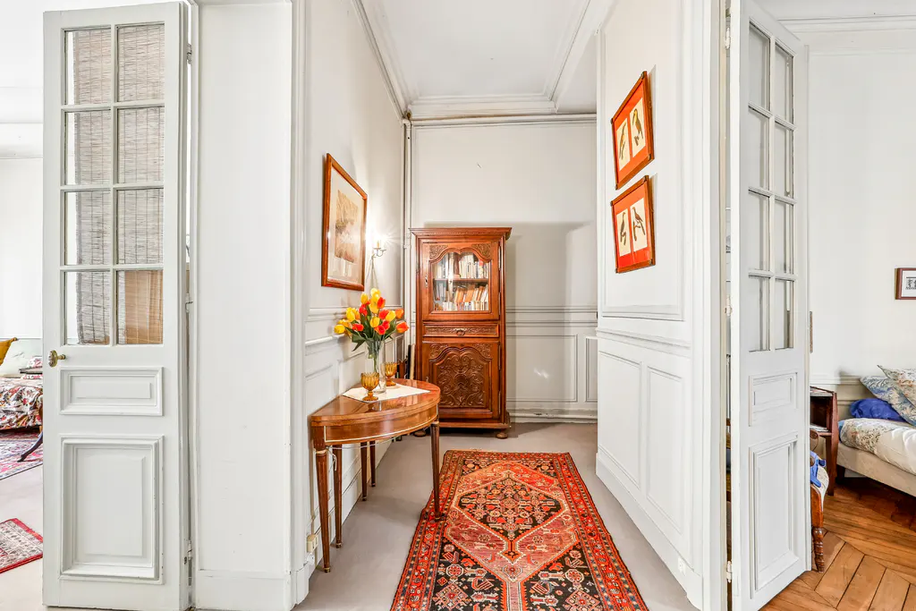 Hallway with white walls, wood floors, and an oriental rug. A wooden cabinet and table with flowers are visible. White paneled doors are on either side.