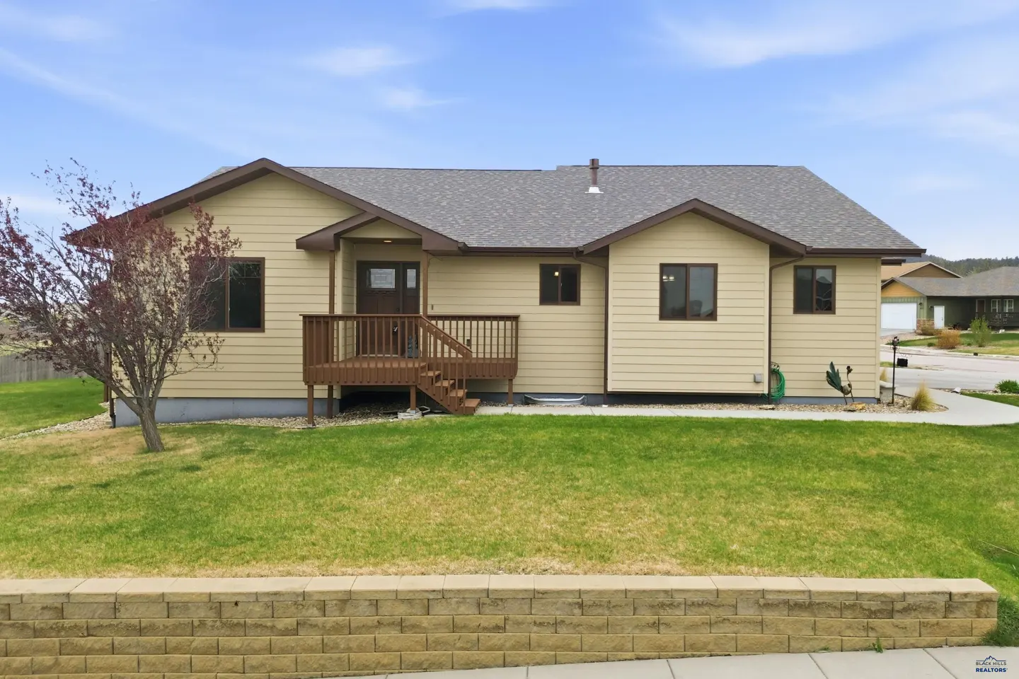 Tan single-story house with a brown deck, gray roof, and green lawn under a blue sky.