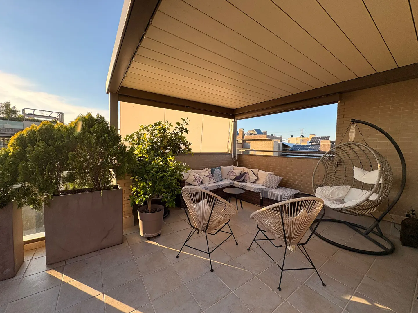 Covered rooftop terrace with modern patio furniture, potted plants, and a hanging egg chair at sunset.