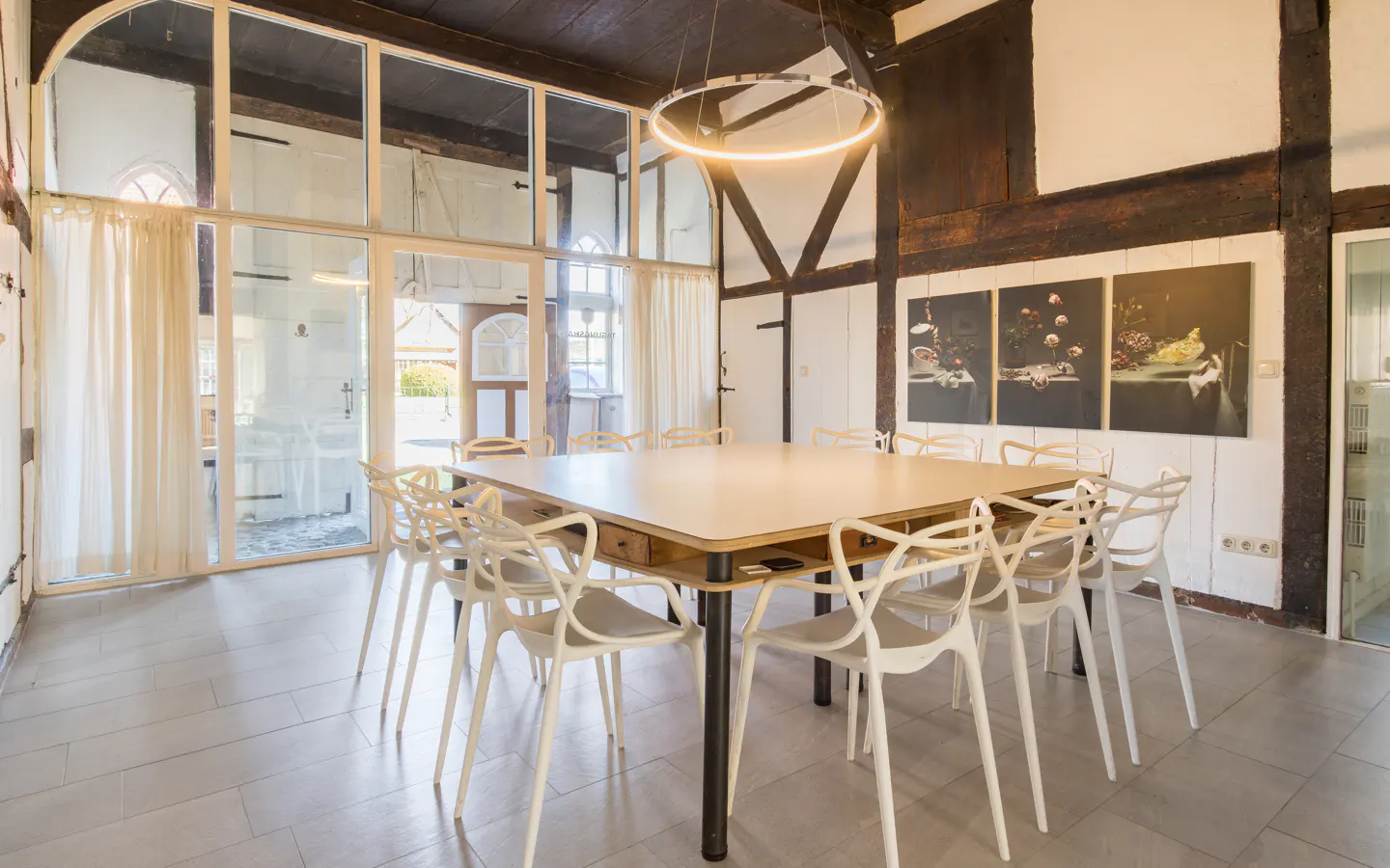Bright room with a large square table surrounded by white chairs. A modern ring light hangs above. Large windows and dark wood beams add character.