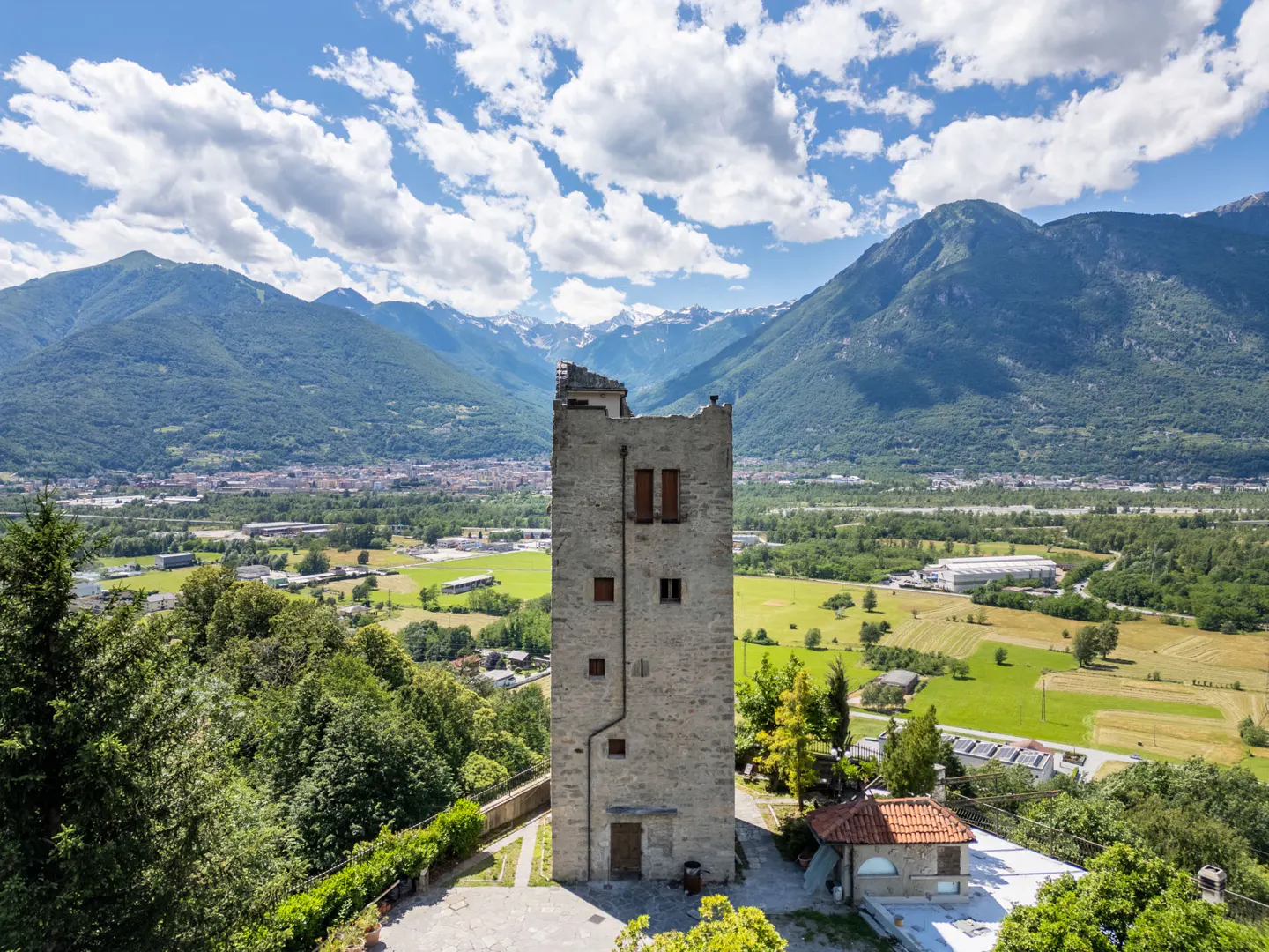 Stone tower with brown shutters, set against a backdrop of green mountains and a blue sky with white clouds.