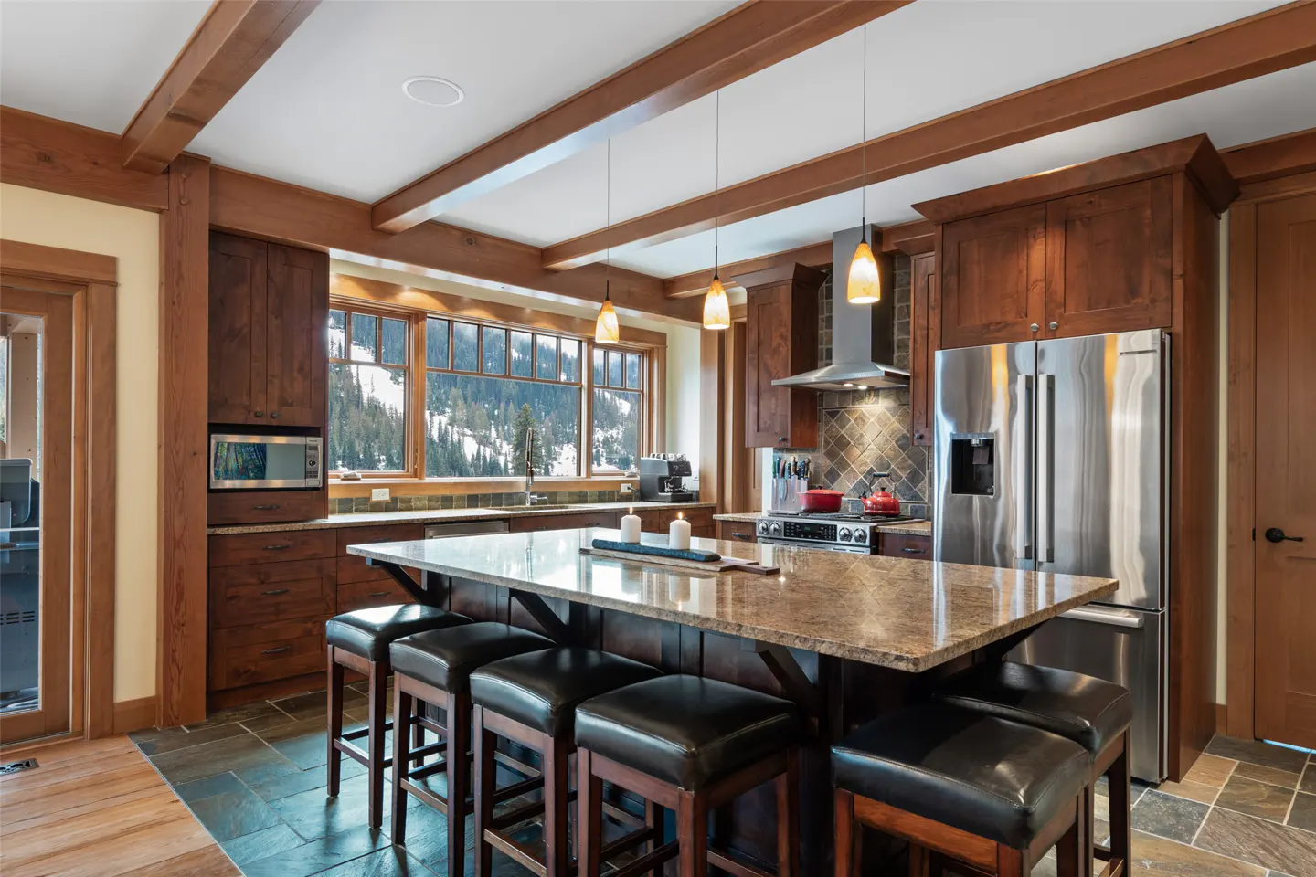 A kitchen with dark wood cabinets, a granite island with black stools, and a view of snow-covered mountains.
