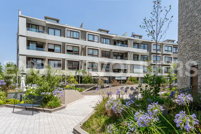 Modern multi-story apartment building with balconies, overlooking a landscaped courtyard with purple flowers and a bench.