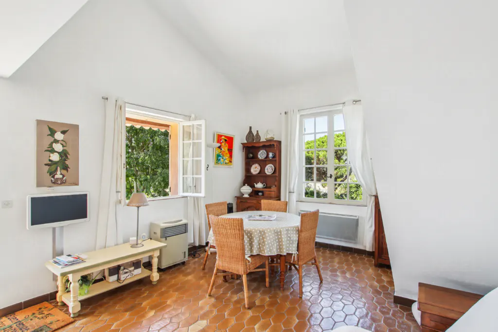 Bright room with white walls, terracotta tile floor, and pitched ceiling. Wicker chairs surround a round table. Windows offer views of greenery.
