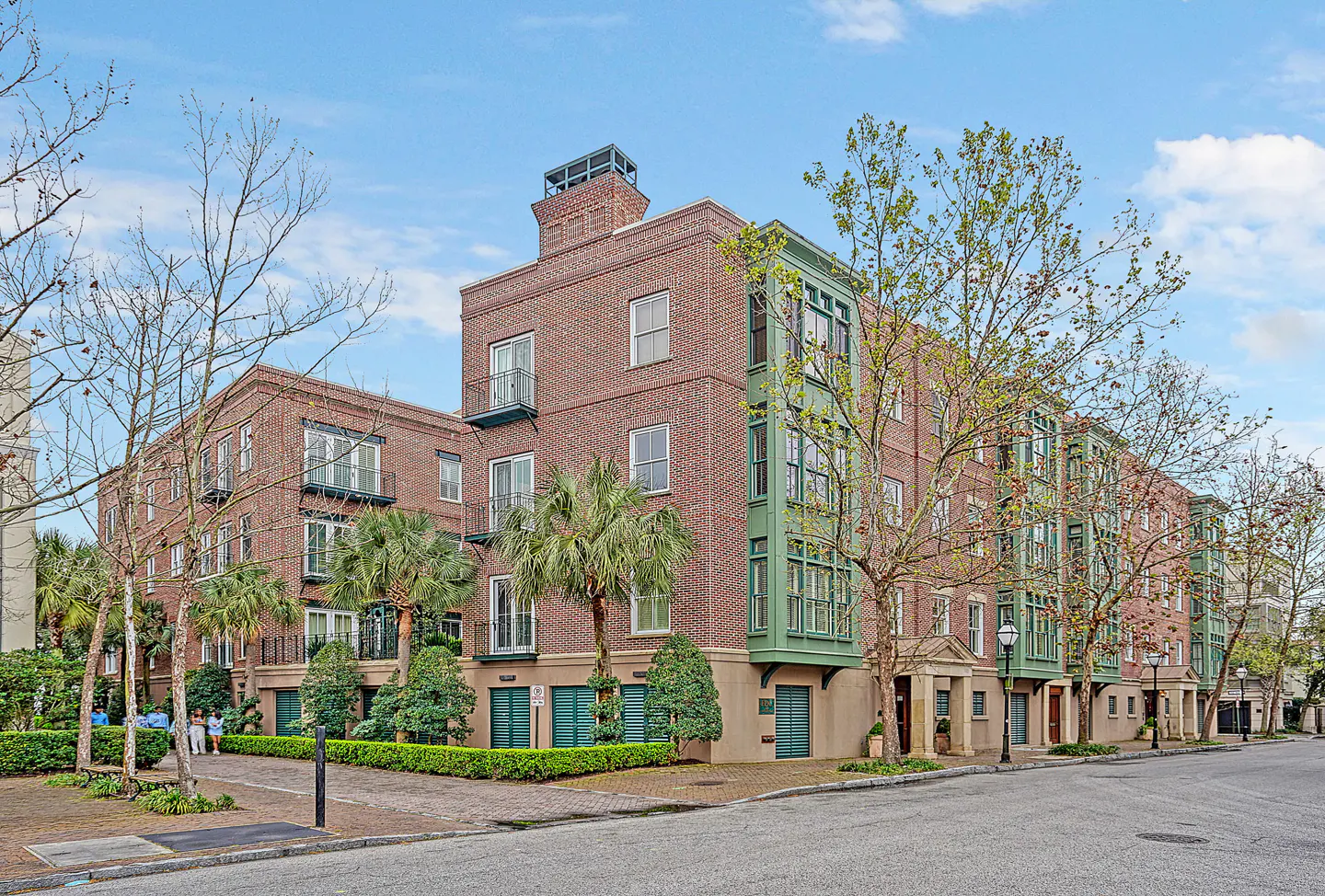 Brick apartment building with green trim, balconies, and palm trees on a sunny day.